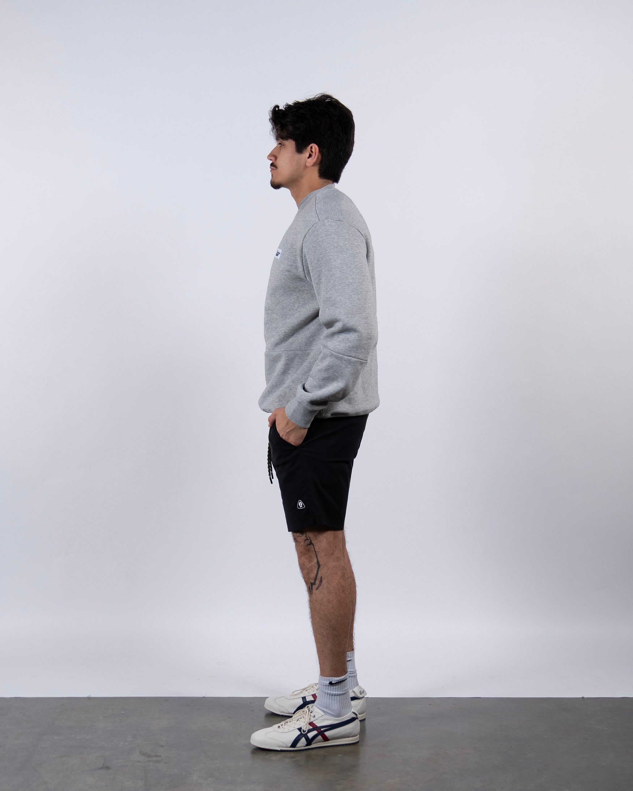 A man stands in profile against a white background, wearing the beardedgoat Men's Void Crew, black shorts, white socks, and white sneakers with black stripes. His hands are in his pockets as he faces left.