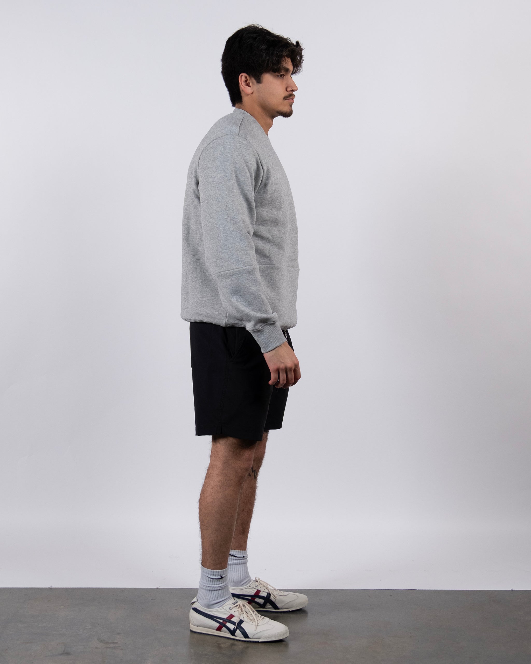 A man stands in profile against a plain background, wearing the beardedgoat Men's Void Crew sweatshirt in gray with ribbed knit cuffs, paired with black shorts, white socks, and white sneakers featuring blue and red stripes.