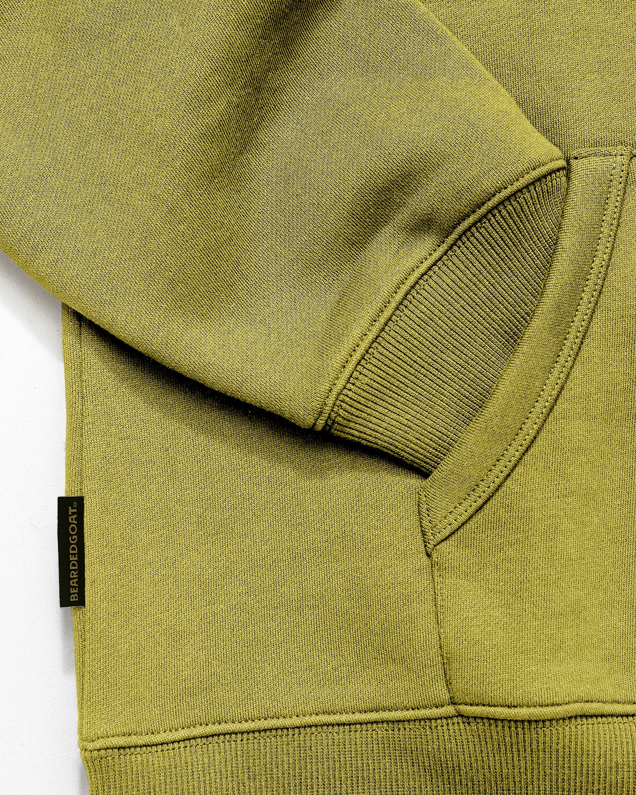 Close-up of The VOID™ Hoodie by beardedgoat, a men's green heavyweight cotton fleece hoodie, showing the ribbed cuff, part of the pocket, and a small black fabric tag with yellow BARDOOCA text.