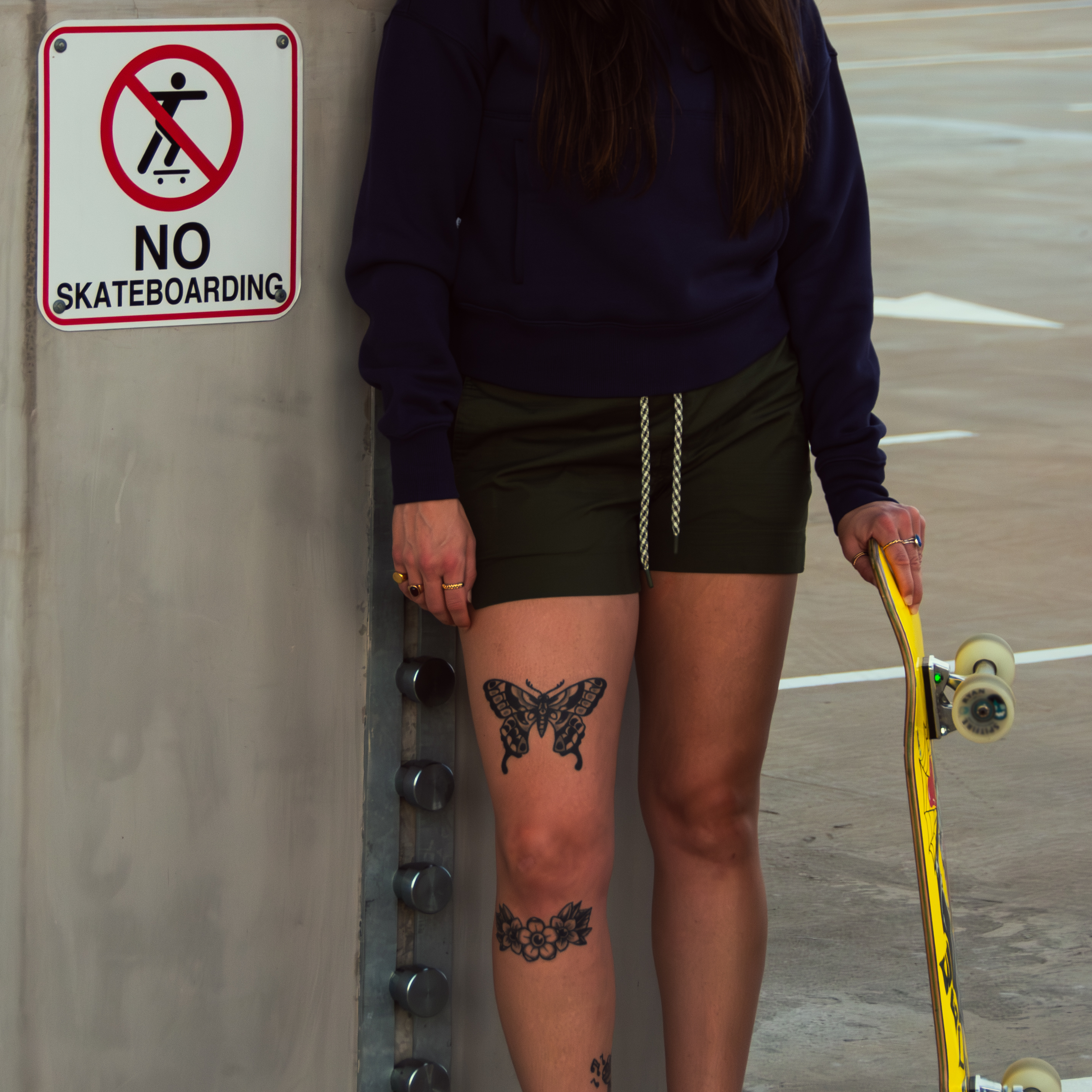 A person with a skateboard stands by a No Skateboarding sign in a parking garage, wearing BEARDEDGOAT’s Women's Canopy Short and a navy sweatshirt, with visible tattoos on their thigh and knee.