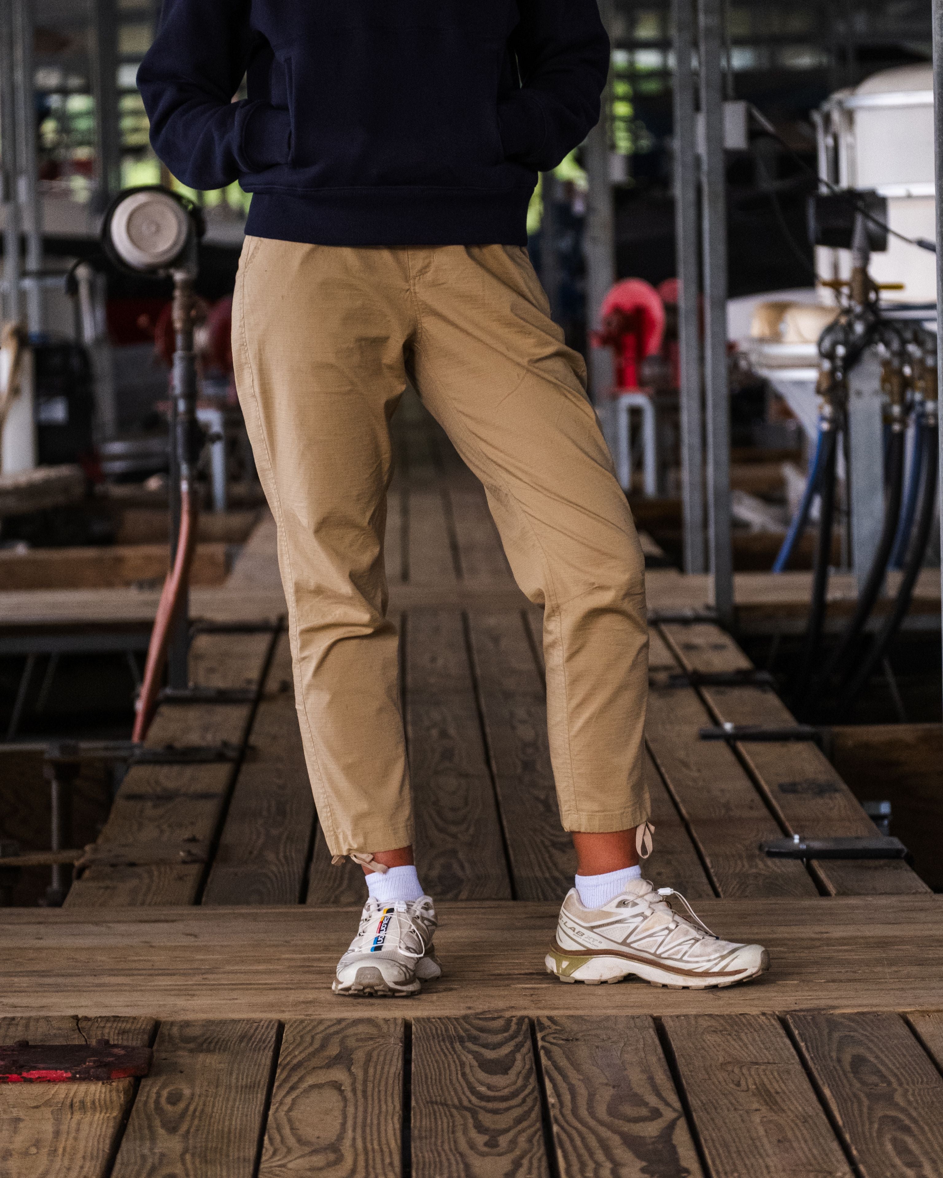 A woman stands on a wooden walkway in BEARDEDGOAT Women's Canopy™ Pant—tan adventure-ready pants—paired with a dark sweatshirt and white sneakers; her upper body is out of frame, with industrial equipment and pipes in the background.