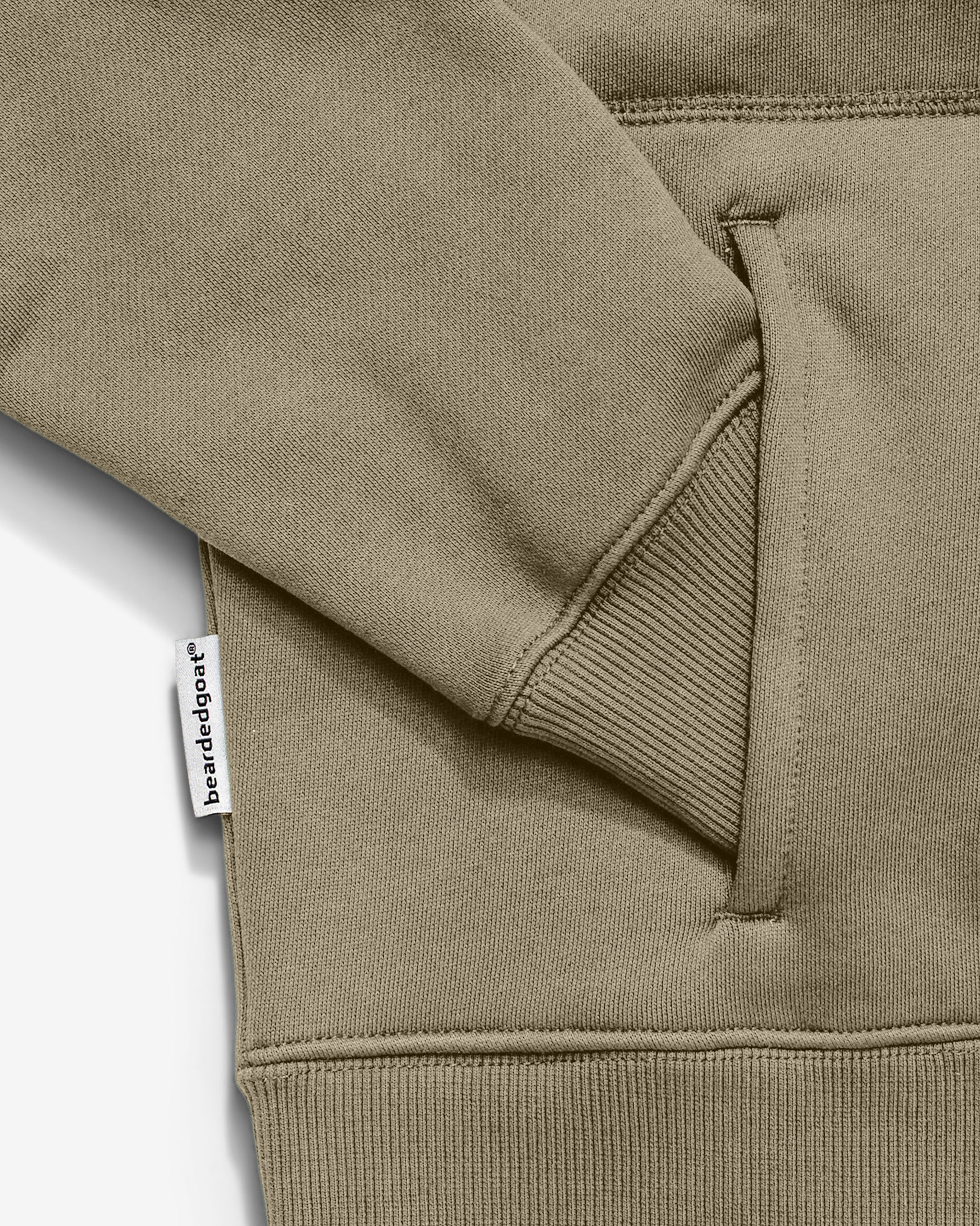 Close-up of the khaki NEW — VOID™ Half Zip by BEARDEDGOAT shows a heavyweight fleece pocket with a ribbed cuffed sleeve resting on it and a white "BEARDEDGOAT" tag sewn into the side seam near the pocket.