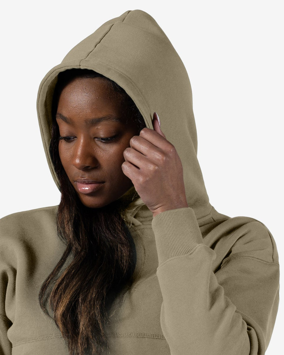 A woman wears the beardedgoat Women's Overnight™ Hoodie in light olive green with matching sweatpants, standing against a plain white background, looking down and adjusting her hood.