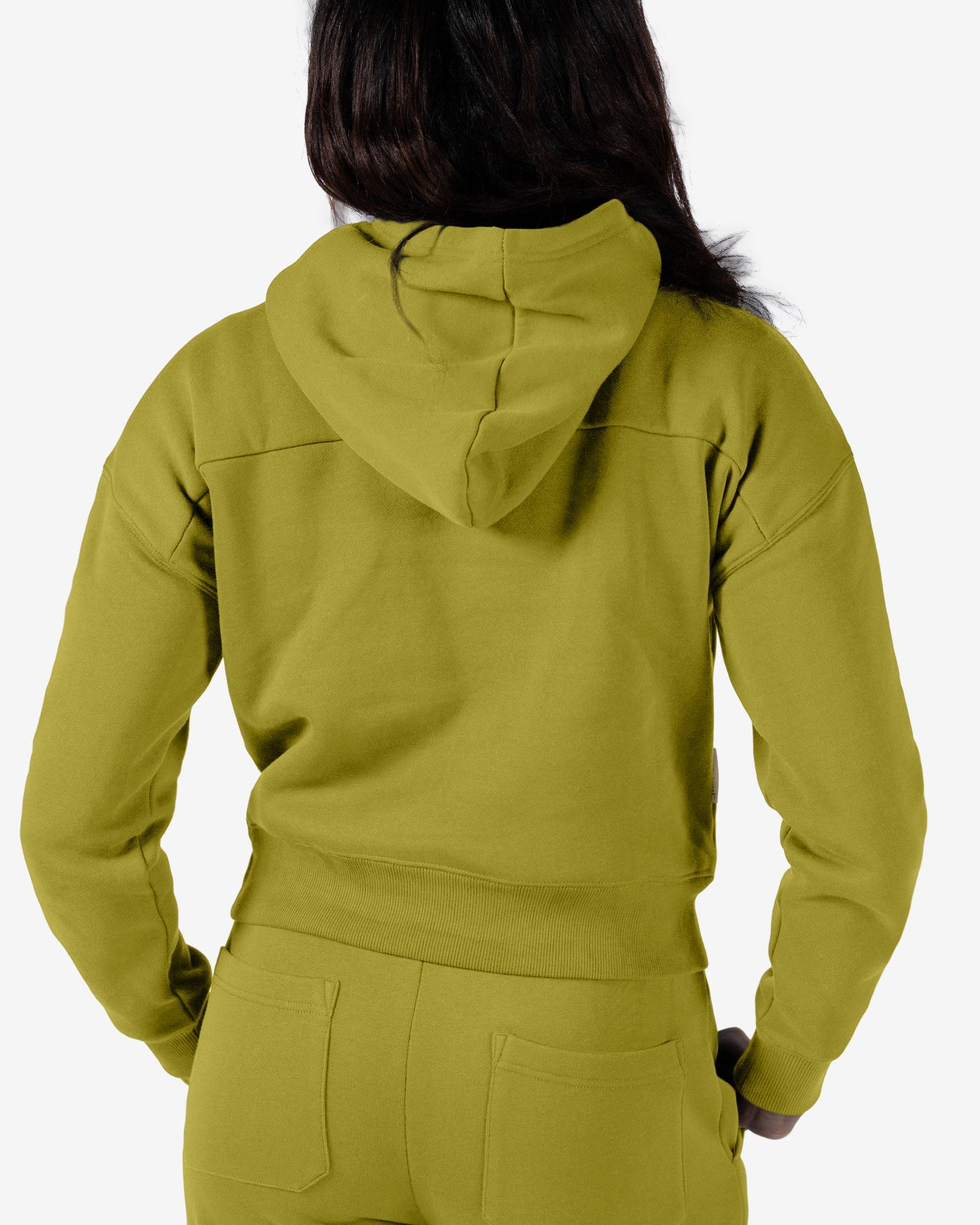 A woman with long dark hair is shown from the back in a matching olive-green Women's Overnight™ Hoodie and sweatpants set by beardedgoat, made of heavyweight cotton fleece. The hood is up but not on her head, against a plain white background.