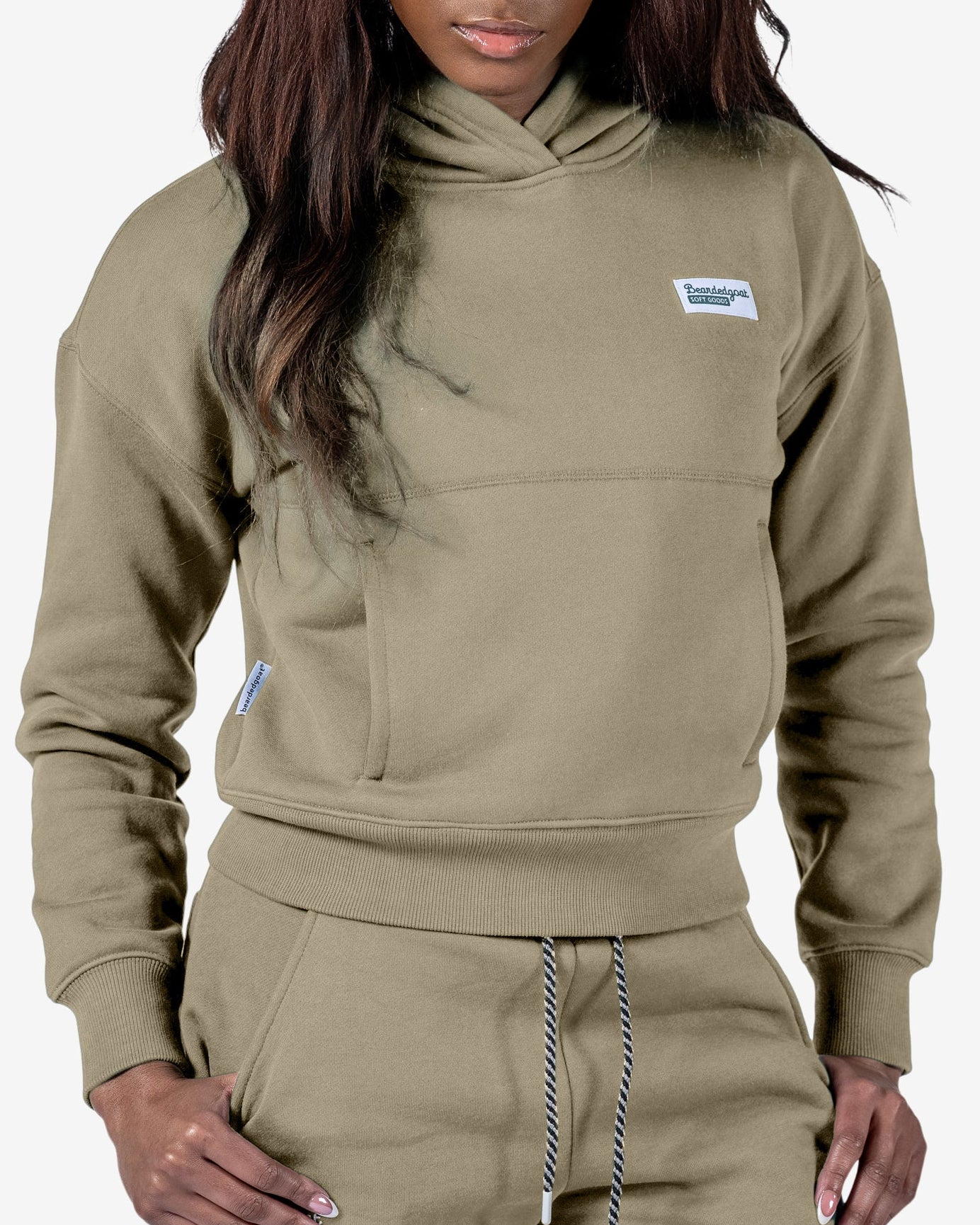 A woman models the beardedgoat Women's Overnight™ Hoodie in tan with a relaxed fit and matching joggers, one hand in her pocket, looking down. The hoodie has a small rectangular patch on the left chest. The background is plain white.