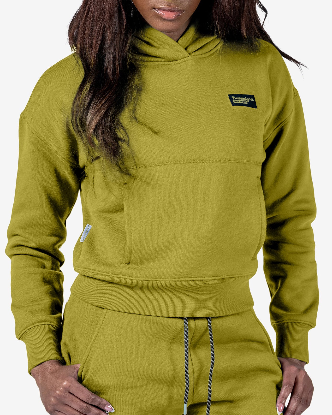 A woman wears the beardedgoat Women's Overnight™ Hoodie and matching sweatpants in olive green, made from heavyweight cotton fleece with a front pouch pocket and a small black rectangular chest patch, creating a casual, sporty look.