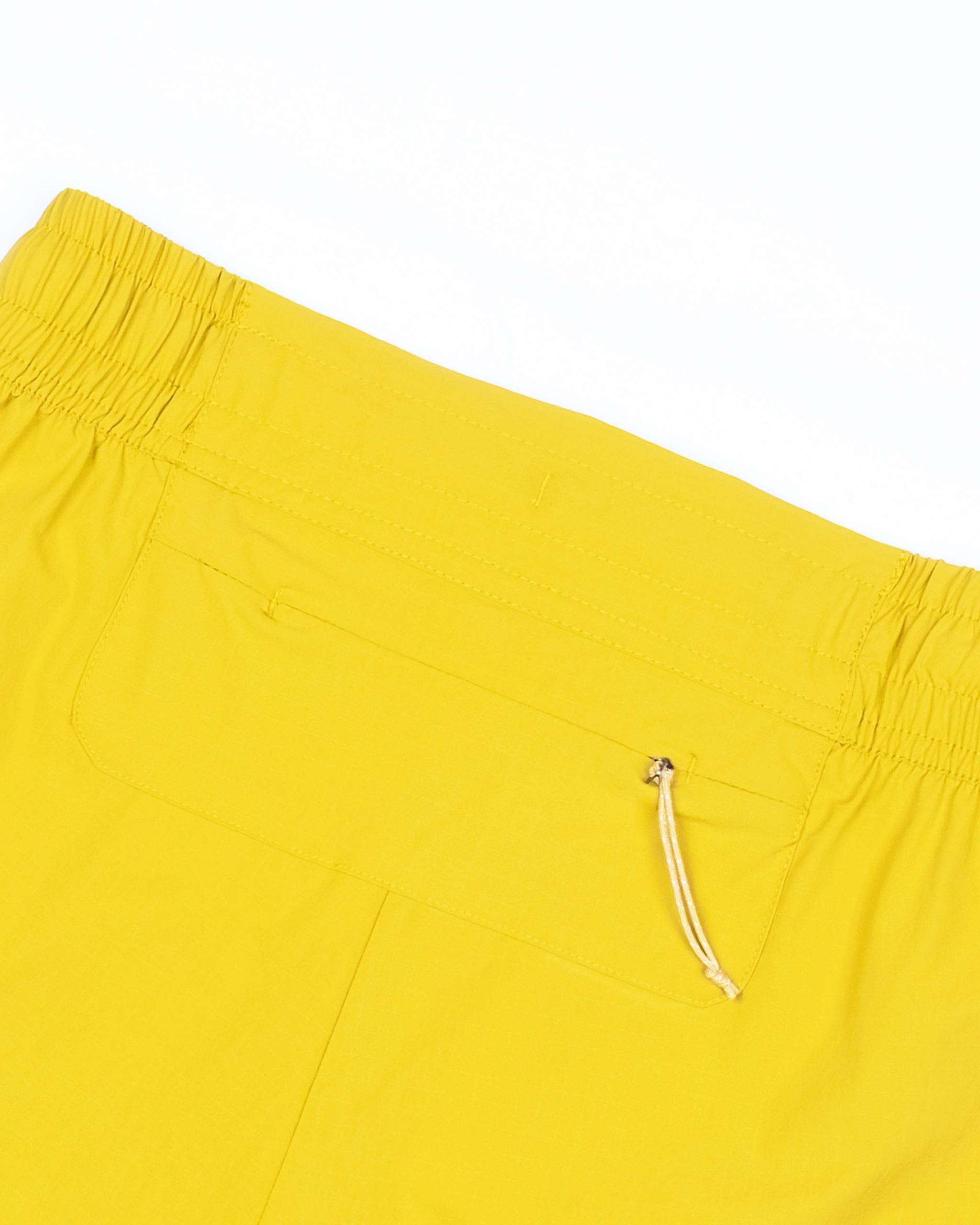 Close-up of the back of BEARDEDGOAT Women's Alpina Short in bright yellow, featuring a high waist, elastic waistband, zippered pocket with beige pull, and durable ripstop fabric against a white background.
