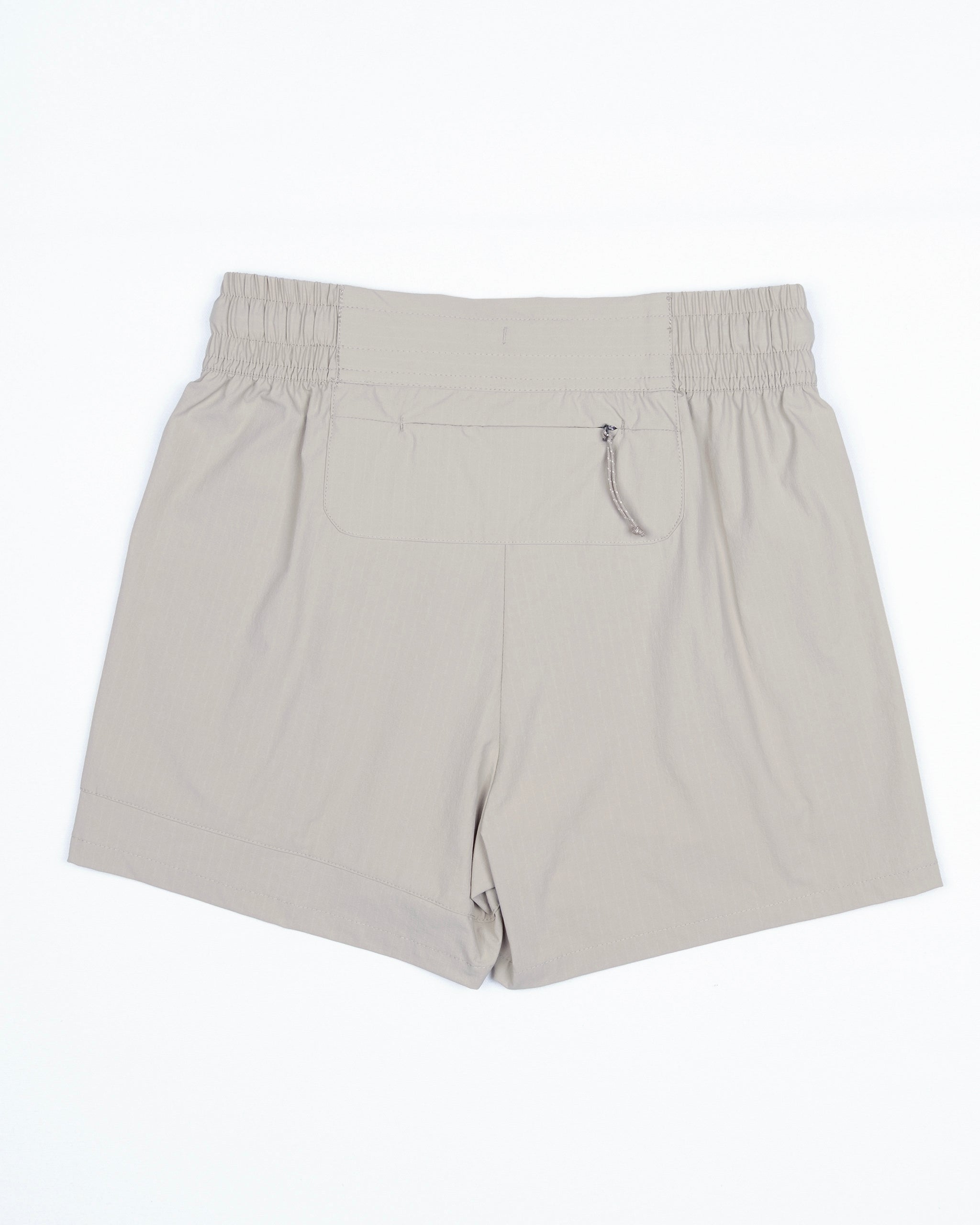 The BEARDEDGOAT Women's Alpina Short in beige is shown flat on a white background, featuring an elastic waistband with gathered details, a back pocket, zippered pockets, and a pull tab.