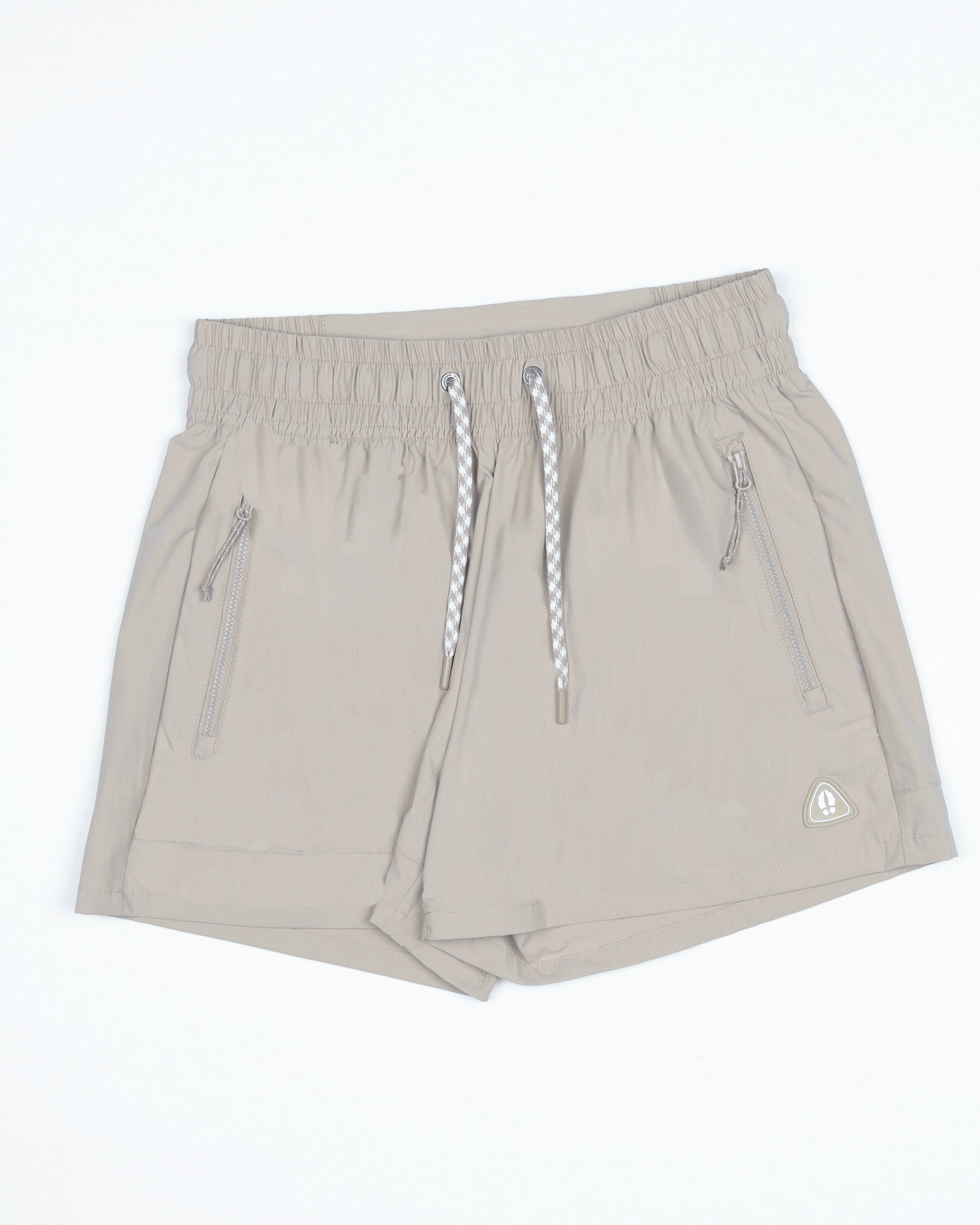The Women's Alpina Short by BEARDEDGOAT features light beige durable ripstop fabric, an elastic waistband with white and gray drawstrings, two zippered side pockets, and a small logo near the bottom left hem.