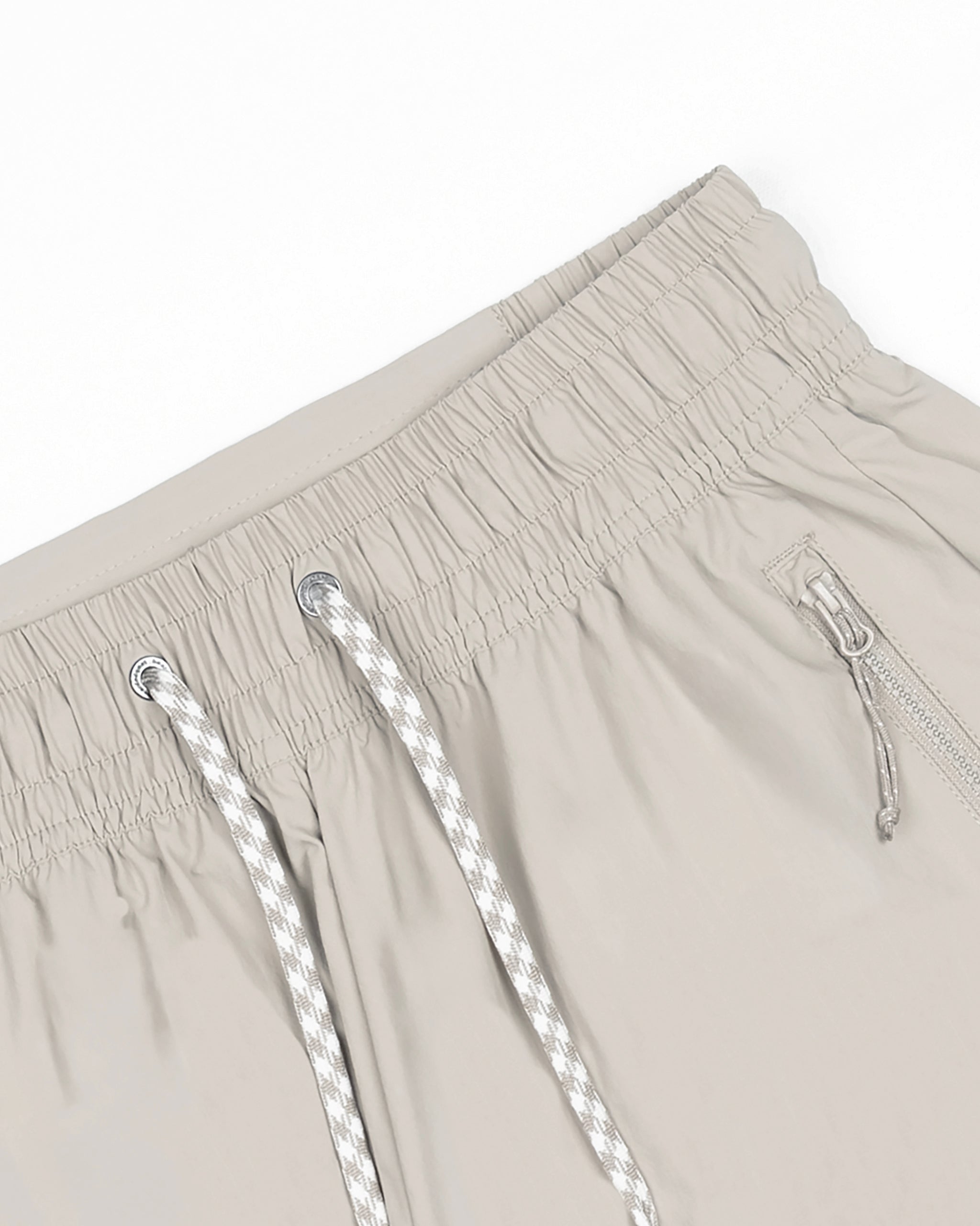 The BEARDEDGOAT Women's Alpina Short in beige features an elastic waistband with drawstring eyelets, a right zippered pocket, and durable ripstop fabric. Shown on a white background.