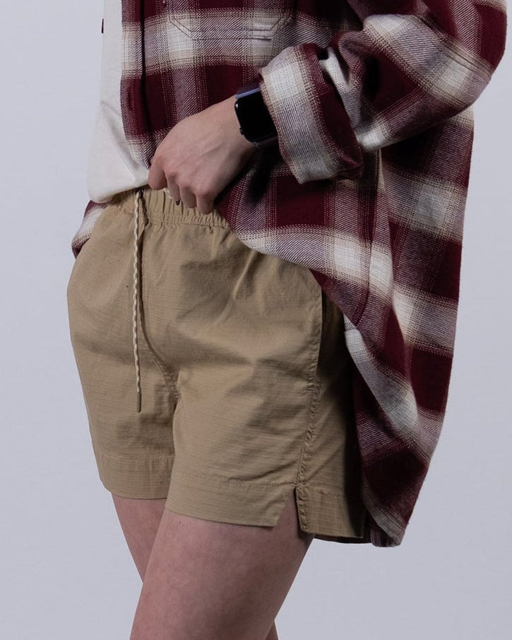 A woman with long dark hair, in profile against a plain background, wears BEARDEDGOAT Women's Canopy Shorts with stretch cotton ripstop, a red and white plaid shirt, gray socks, and white sneakers, looking slightly forward.
