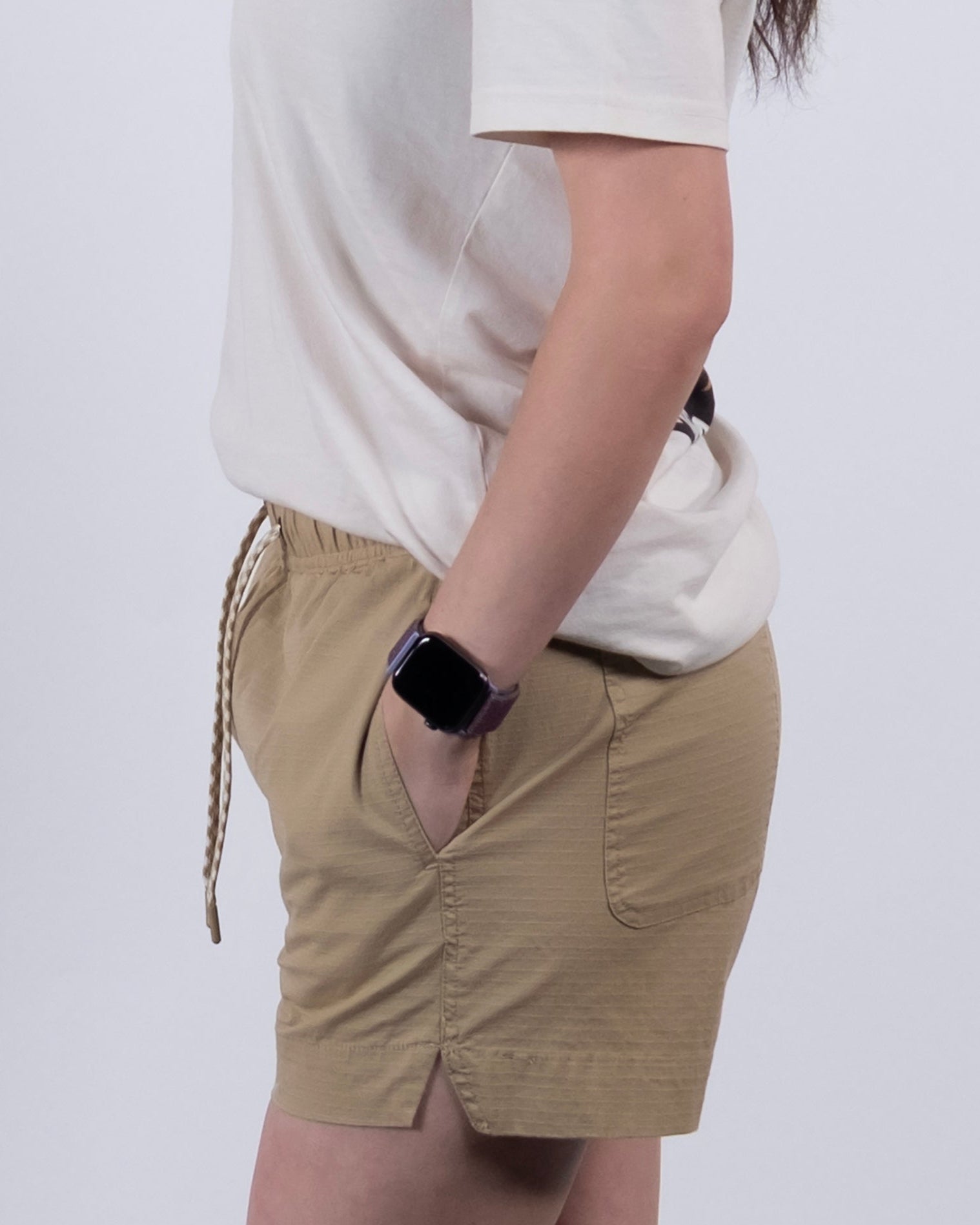 A person stands sideways against a white background, wearing a white t-shirt, BEARDEDGOAT Women's Canopy Short in beige stretch cotton ripstop with pockets, and a smartwatch on their wrist.