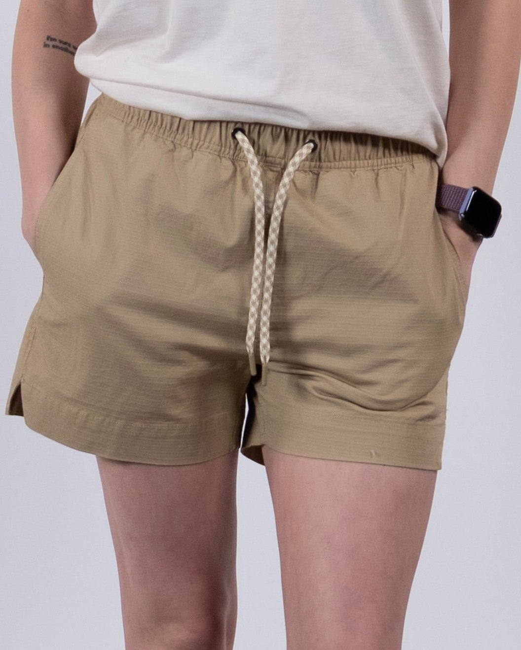 A person stands against a plain background wearing BEARDEDGOAT Women's Canopy Short in beige stretch cotton ripstop, a white t-shirt, frilled white socks, white sneakers, and a smartwatch on their left wrist. Their face is not shown.