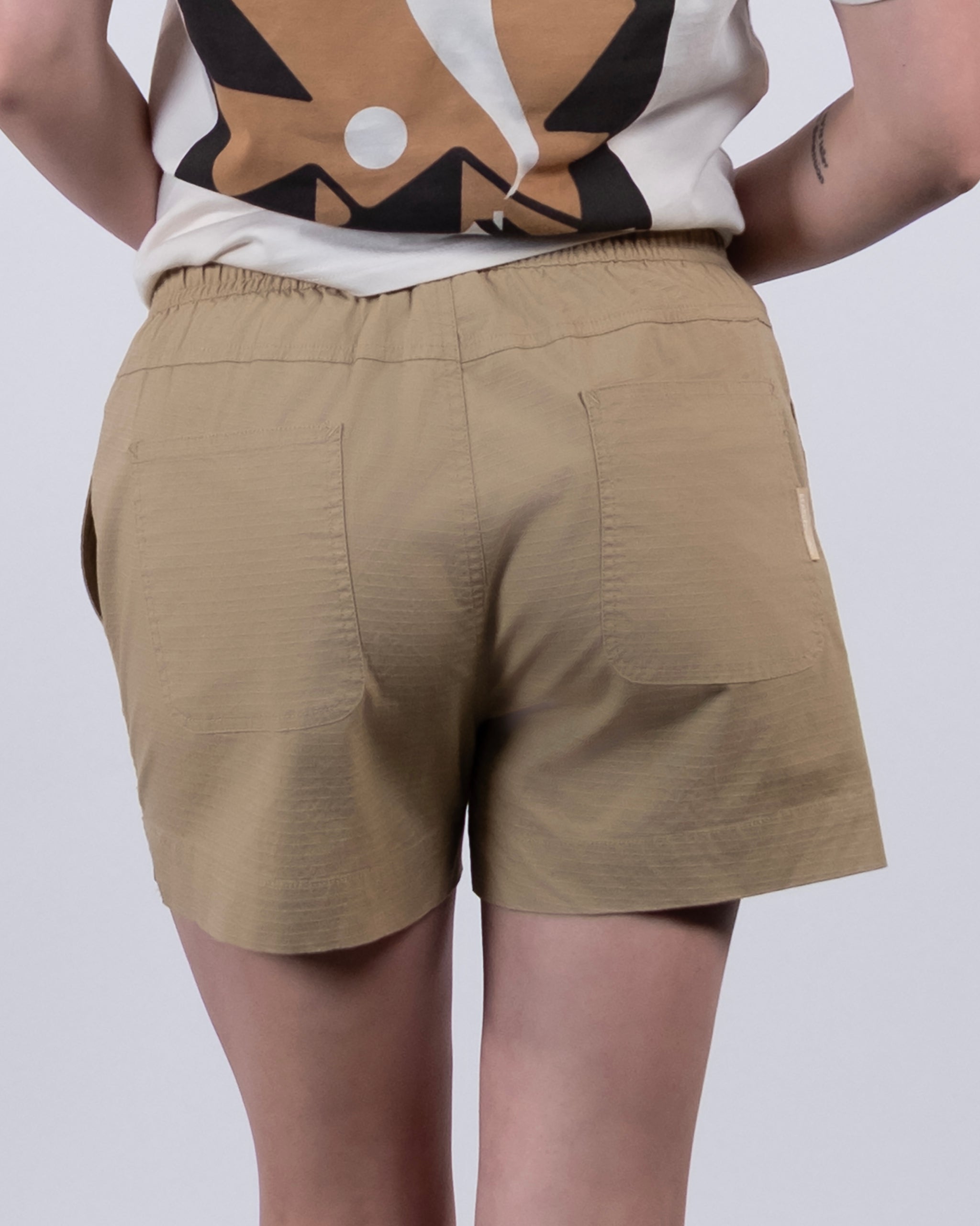 A person is seen from behind wearing BEARDEDGOAT Women's Canopy Shorts in tan with two back pockets and an elastic waistband, paired with a white and brown graphic tee. Their tattooed arm is partly visible.