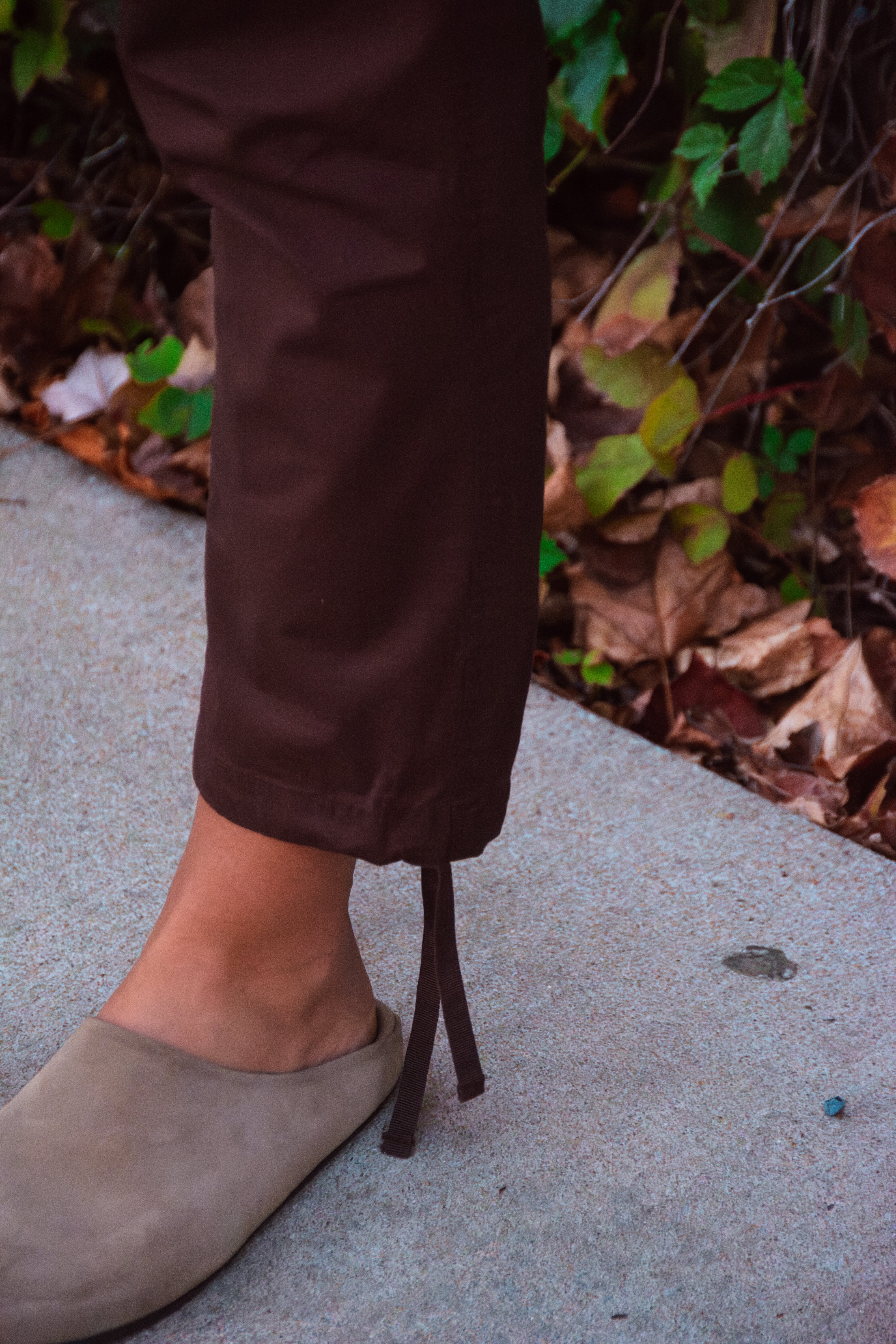 A person stands on a sidewalk near dried leaves and green foliage, wearing BEARDEDGOAT Women's Canopy™ Pant—adventure-ready stretch cotton ripstop pants with ankle drawstrings—and taupe slip-on shoes.