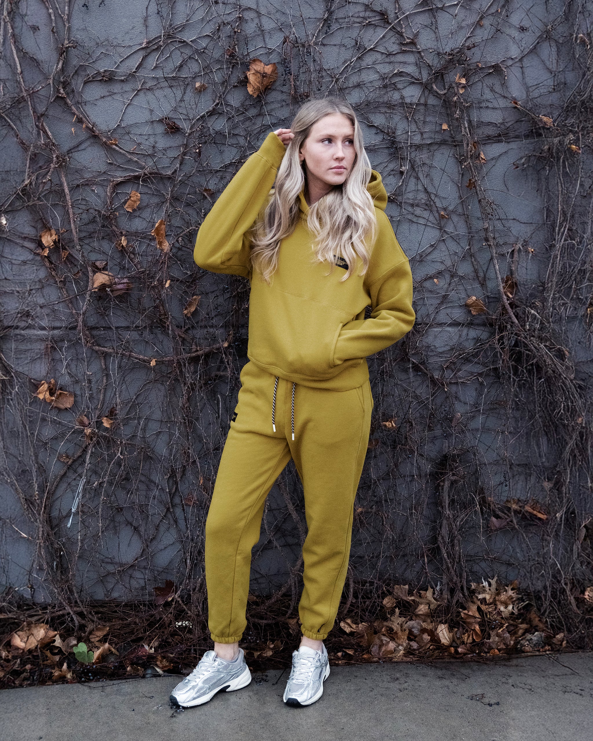 A woman with long blond hair in a beardedgoat Women's Overnight™ Hoodie in mustard yellow, made of heavyweight cotton fleece, stands by a gray vine-covered wall with dry leaves on the ground, wearing white sneakers and touching her hair.