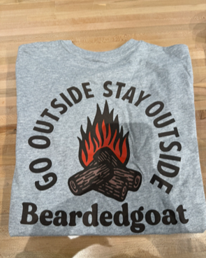 The Campfire Tee - Snow Grey by BEARDEDGOAT is shown folded on a wooden surface, displaying a campfire graphic with the words “GO OUTSIDE STAY OUTSIDE” above and “Beardedgoat” below.