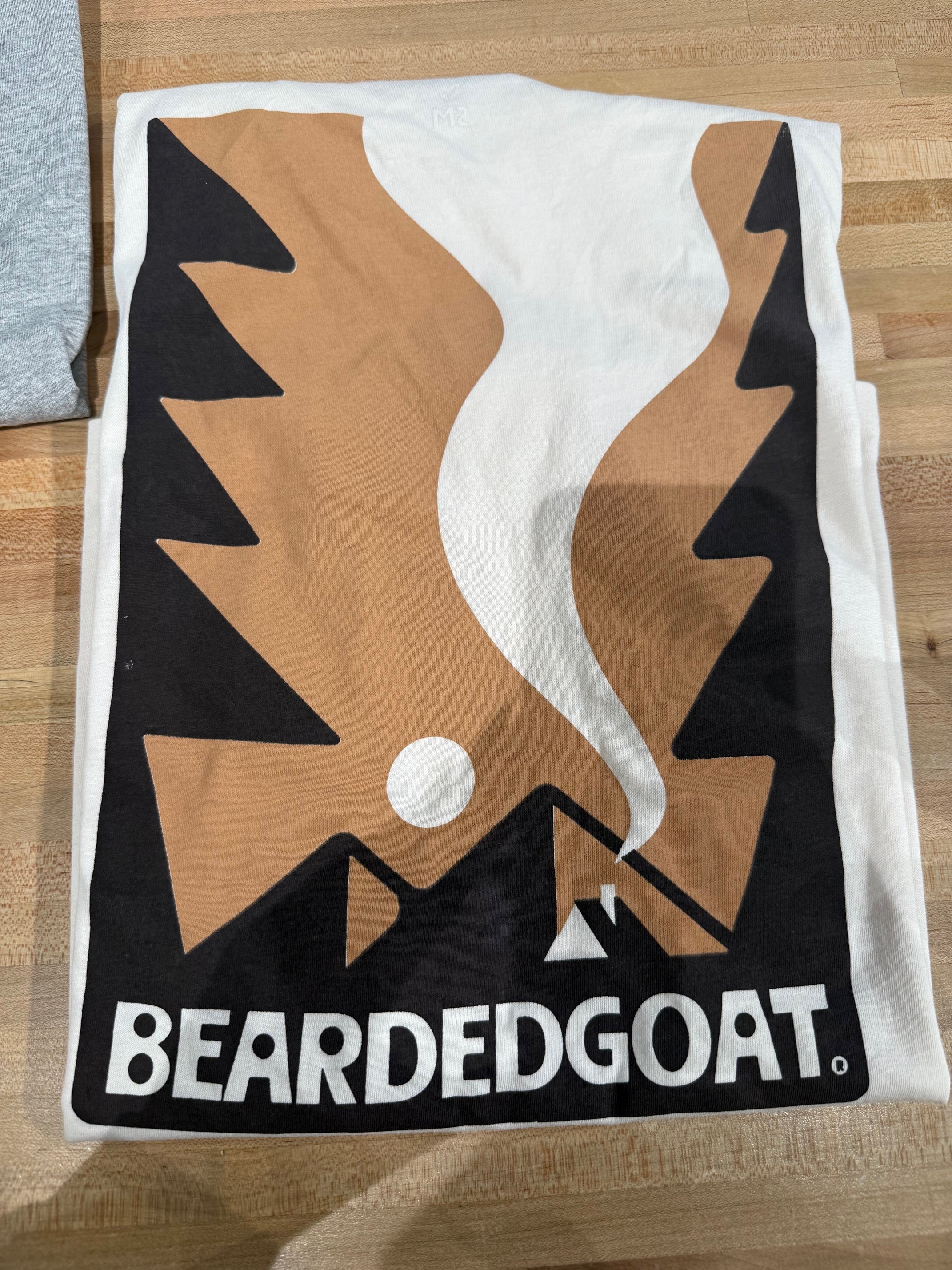 The Valley Camp Tee - Almond by BEARDEDGOAT is a folded white shirt featuring mountain, sun, and campfire graphics with “BEARDEDGOAT” at the bottom, displayed on a wooden surface.