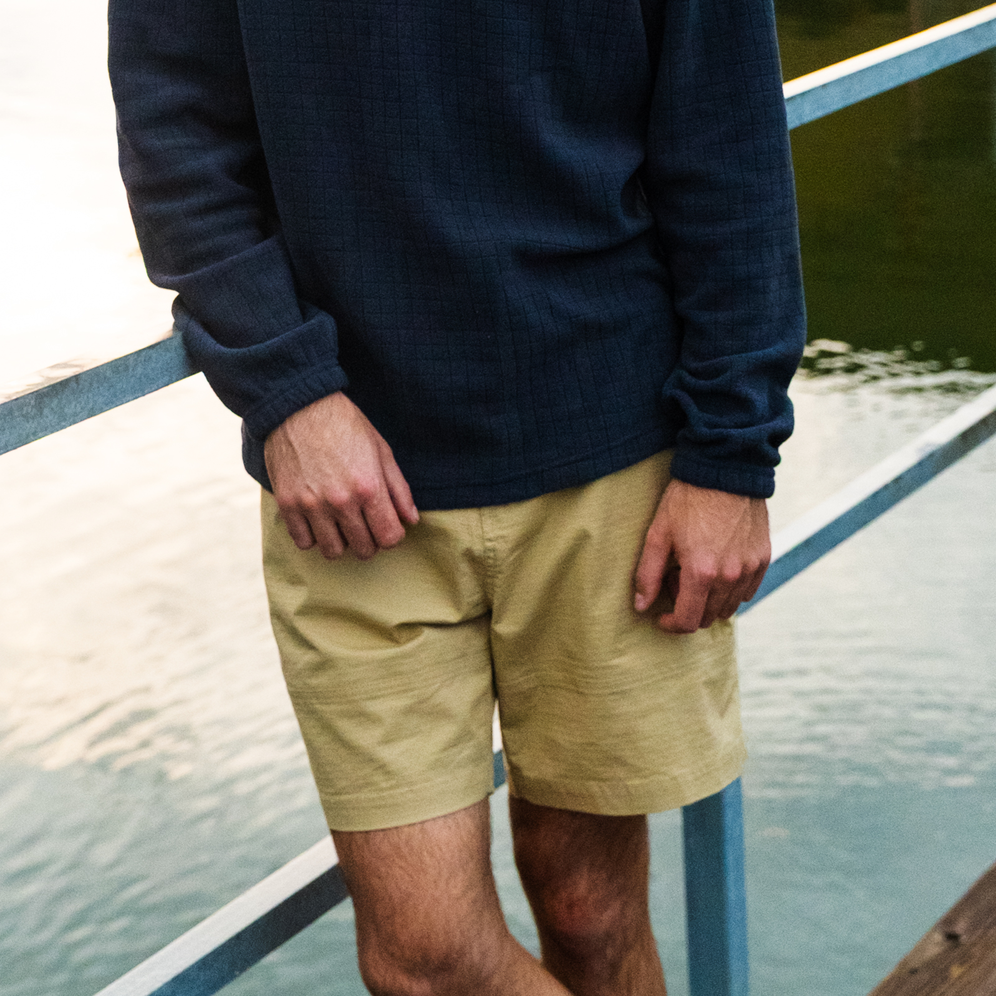 A person in a dark long-sleeve shirt and BEARDEDGOAT Men's Canopy Shorts made from stretch cotton ripstop stands by a metal railing near water, with only their torso and legs visible.