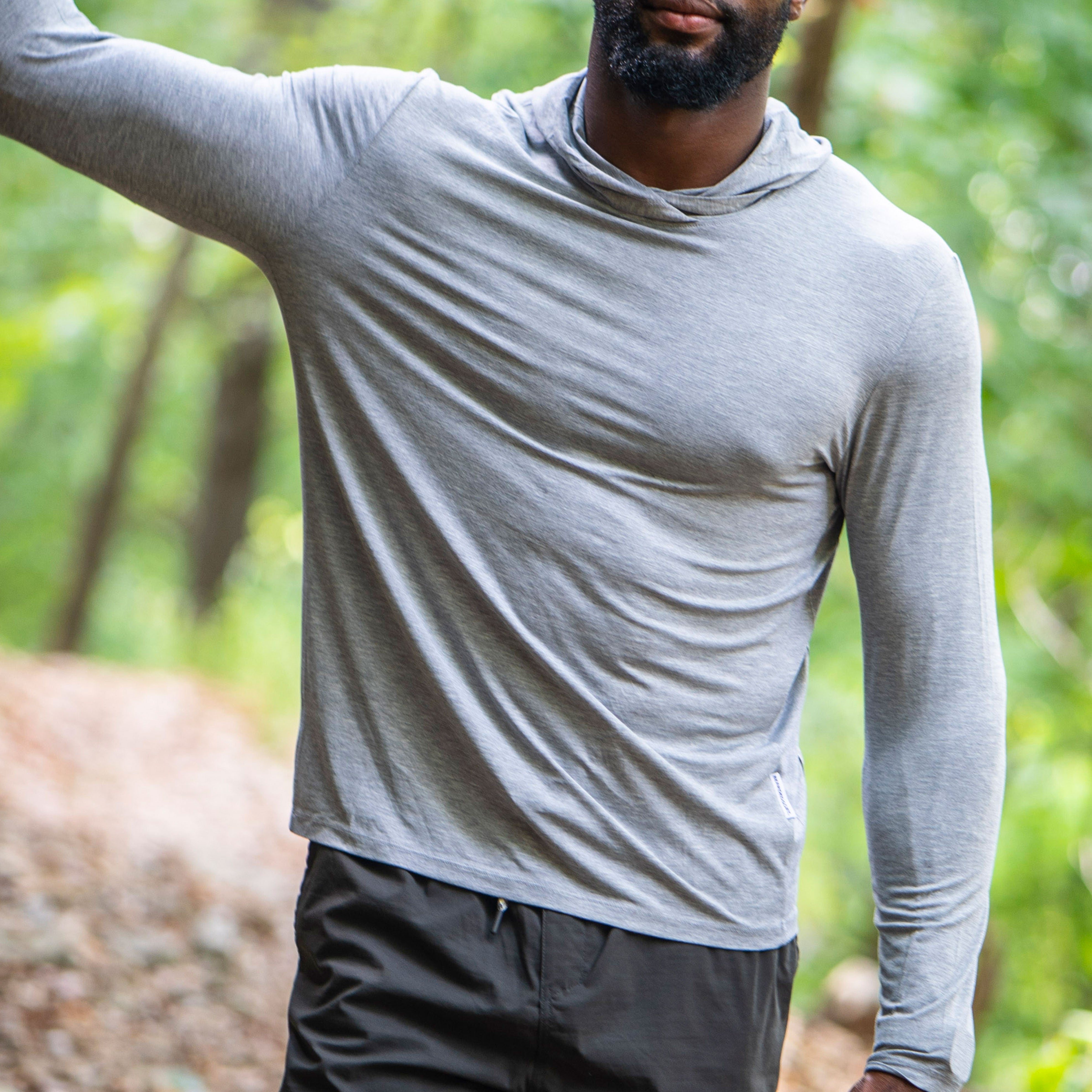 A man stands on a trail among green trees, wearing the BEARDEDGOAT Men’s EverZen™ Hoodie in light gray and black pants, reflecting a commitment to sustainable clothing.
