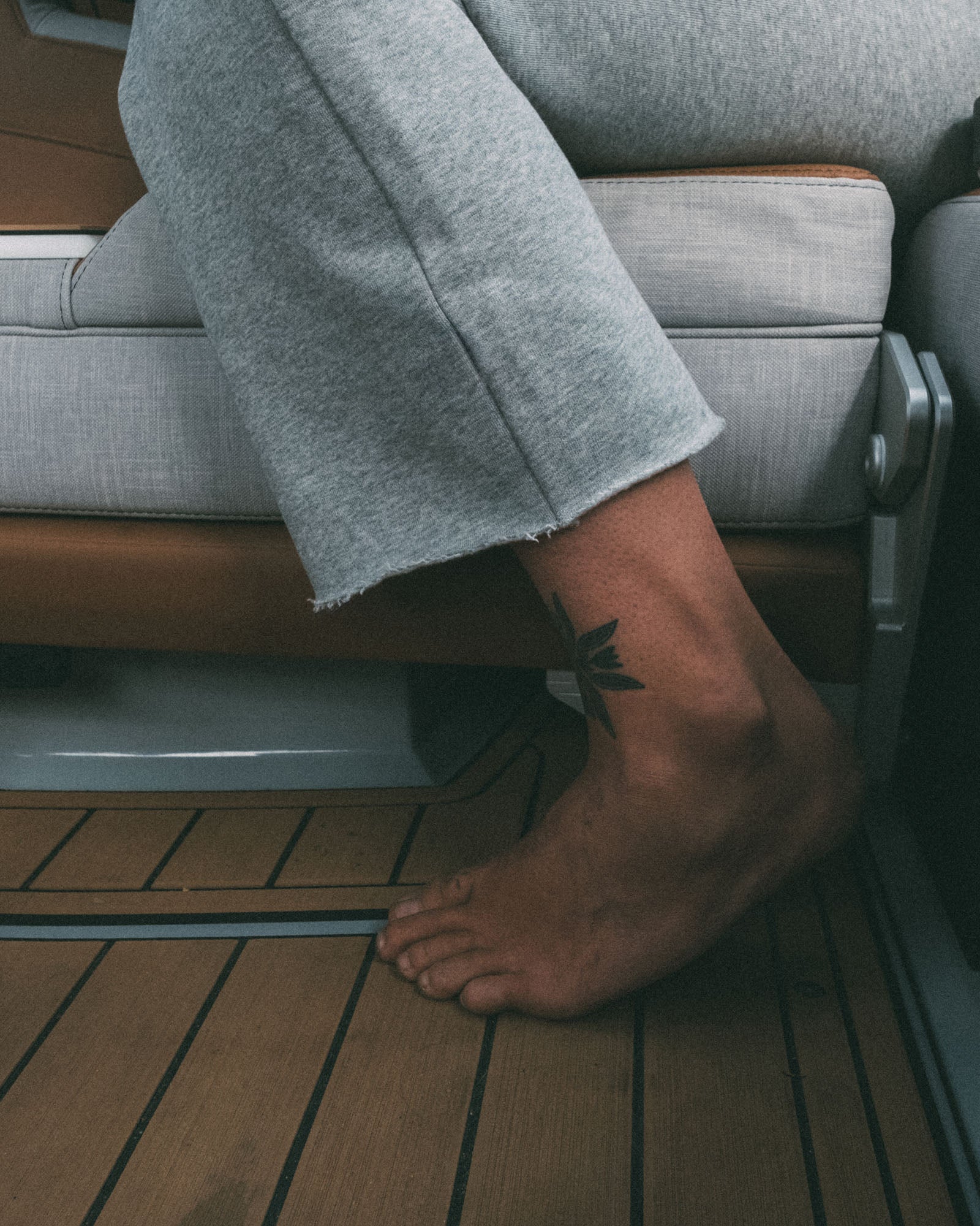 A person wearing beardedgoat’s Women’s Overnight Sweatpant sits on a cushioned seat, with their bare foot on a wooden floor and a black leaf tattoo visible on their ankle.