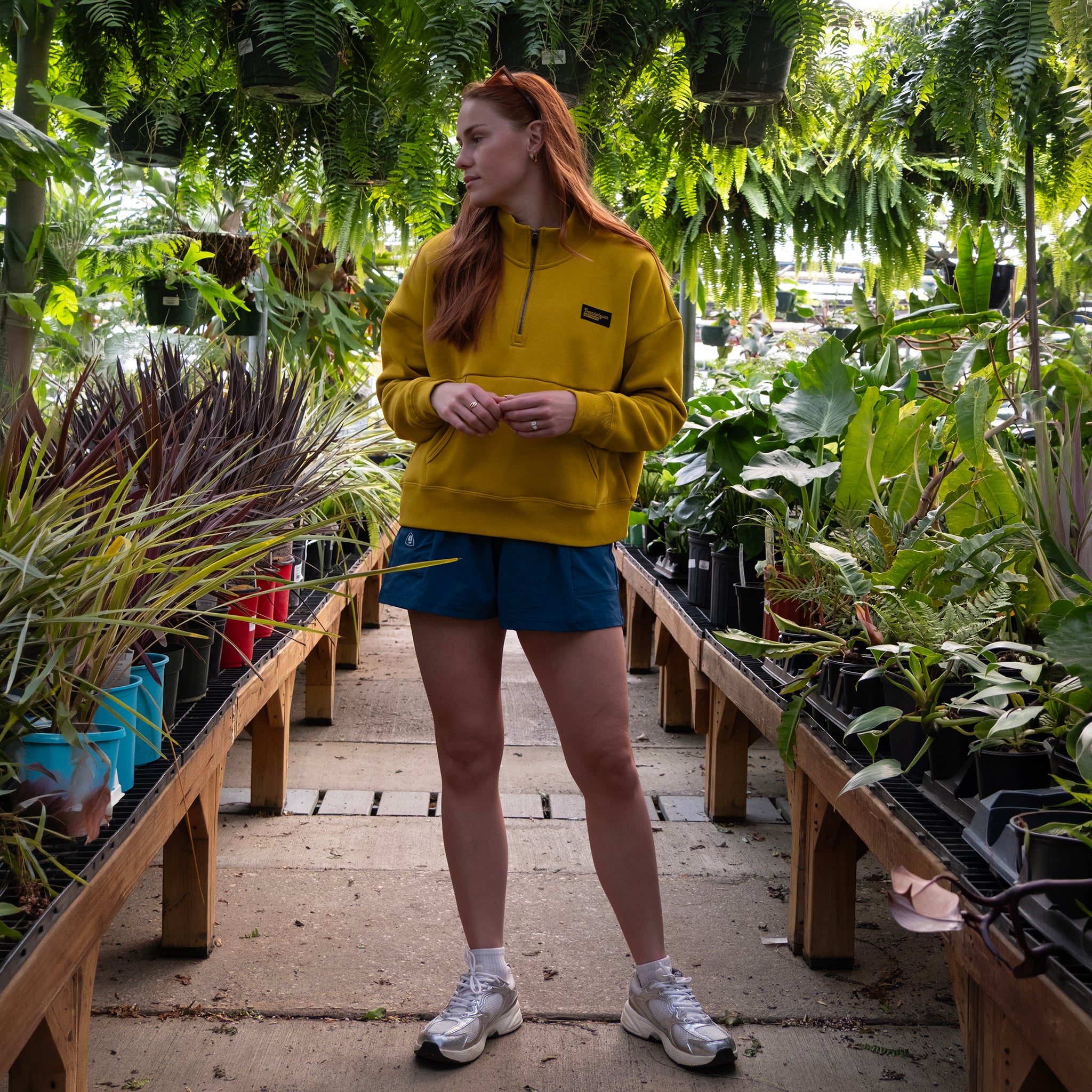 A person with long red hair stands in a plant nursery, wearing the BEARDEDGOAT NEW — Overnight™ Half Zip sweatshirt, blue shorts, and white sneakers, surrounded by green potted plants on wooden tables.