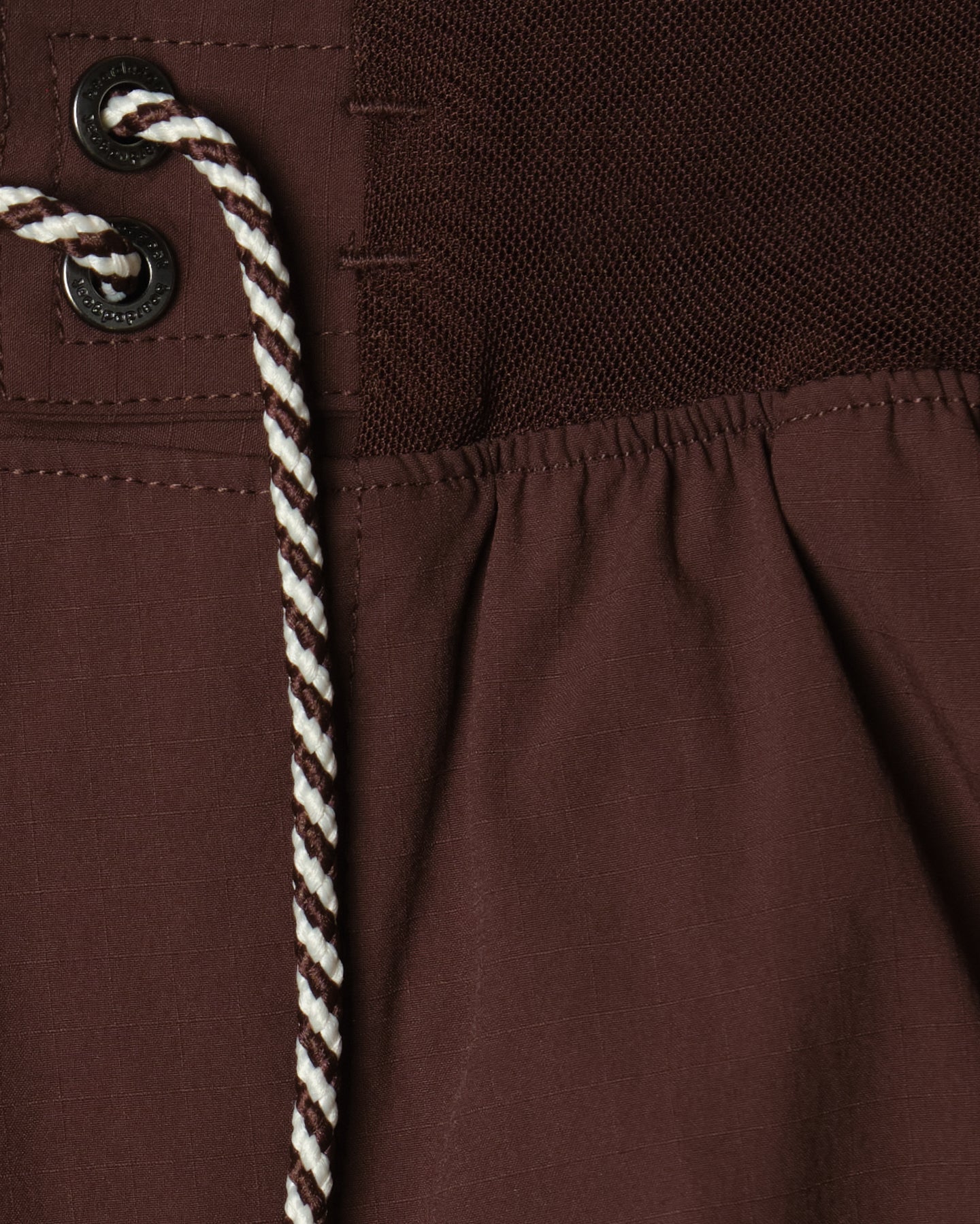 Close-up of BEARDEDGOAT Men's Rambler Short in brown with 4-way stretch, gathered seam, two black metal grommets, white-and-brown braided drawstring, and mesh panel—designed for trail running.