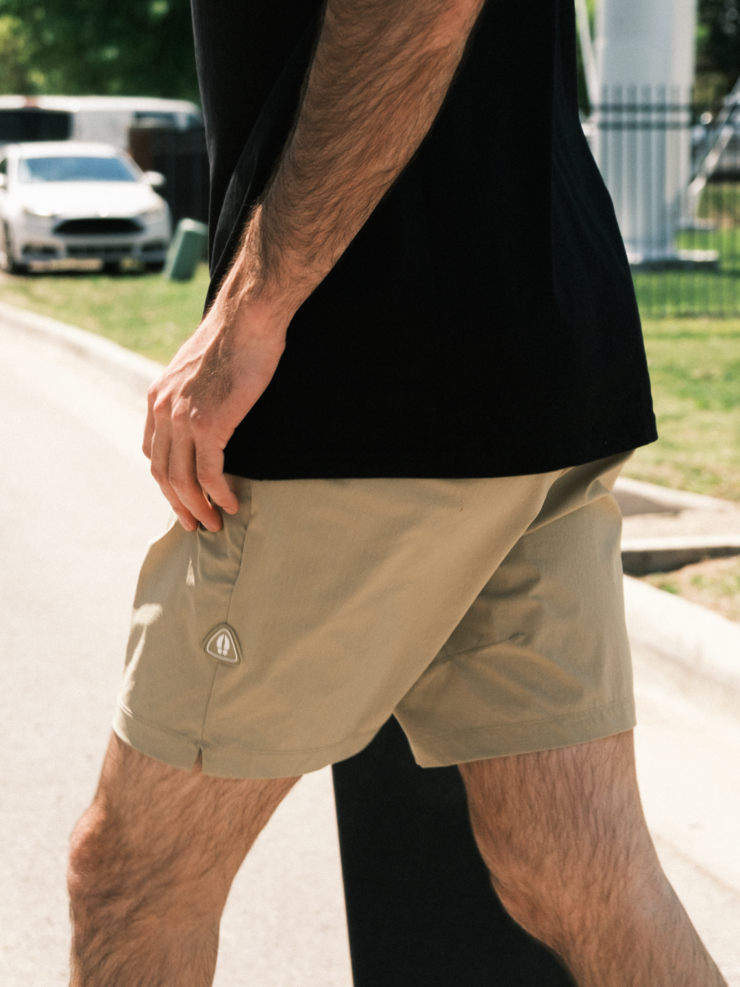 A man wearing BEARDEDGOAT Men's Rambler Short with 4-way stretch is walking outside on a sunny day. Only his lower torso and legs are visible, with a parked car, grass, and a fence in the background.