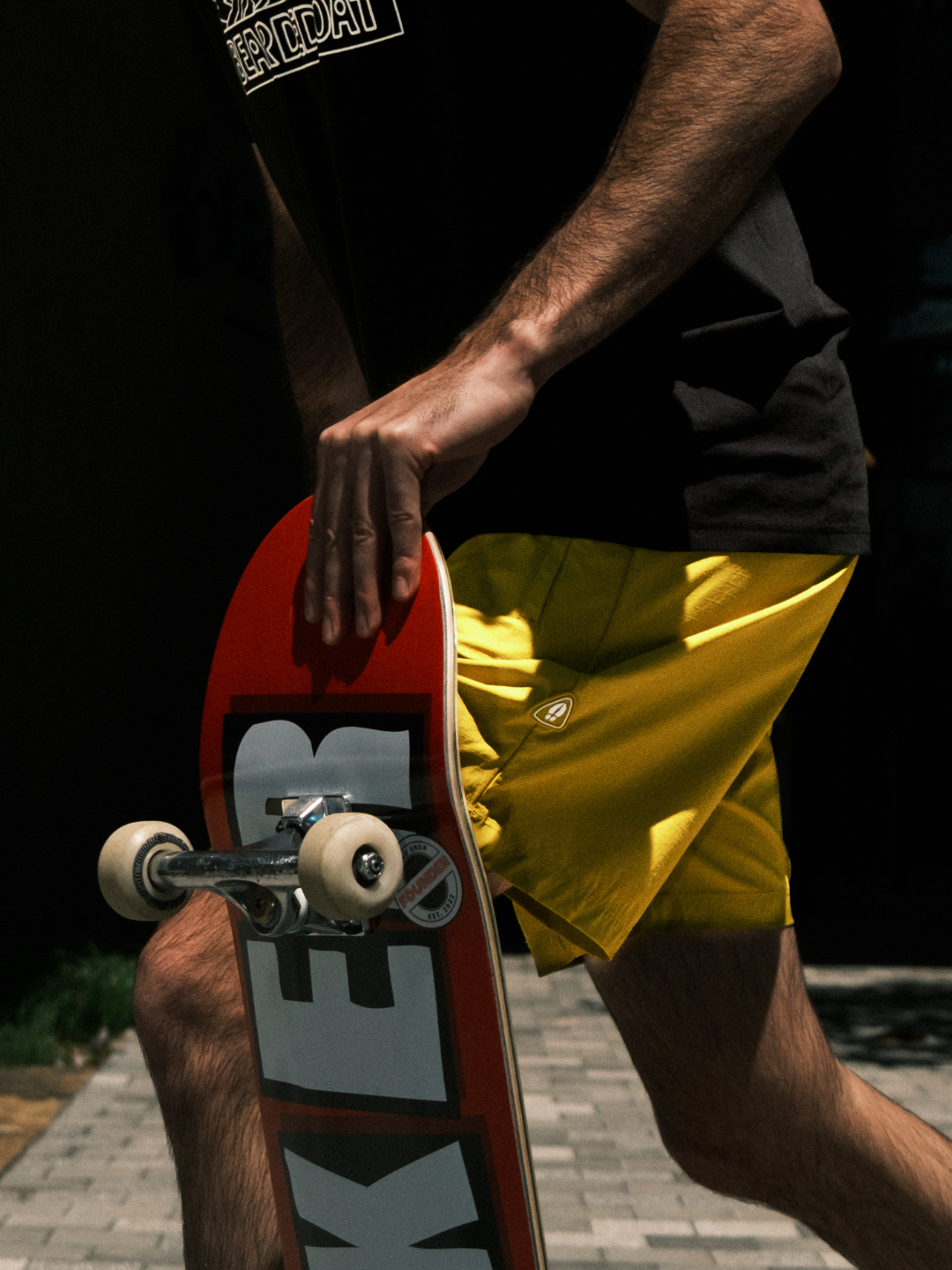 Wearing BEARDEDGOAT's Men's Rambler Short and a black shirt, a person strolls on a sunlit path holding a red skateboard, showing off the 4-way stretch comfort ideal for trail running or laid-back walks.