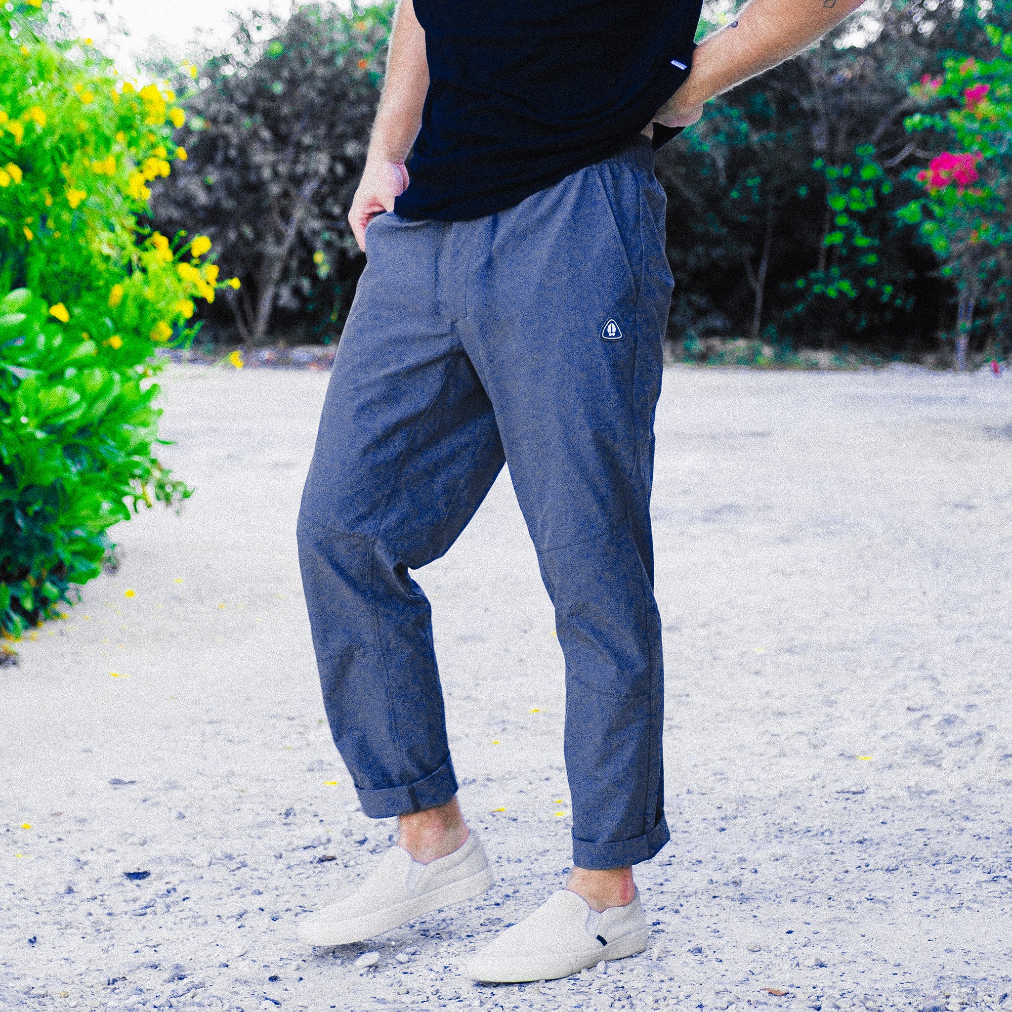 A person wearing BEARDEDGOAT’s NEW — ROVER™ Pant in performance stretch nylon, a black t-shirt, and beige slip-on shoes stands on a gravel path with greenery and yellow and pink flowers in the background.