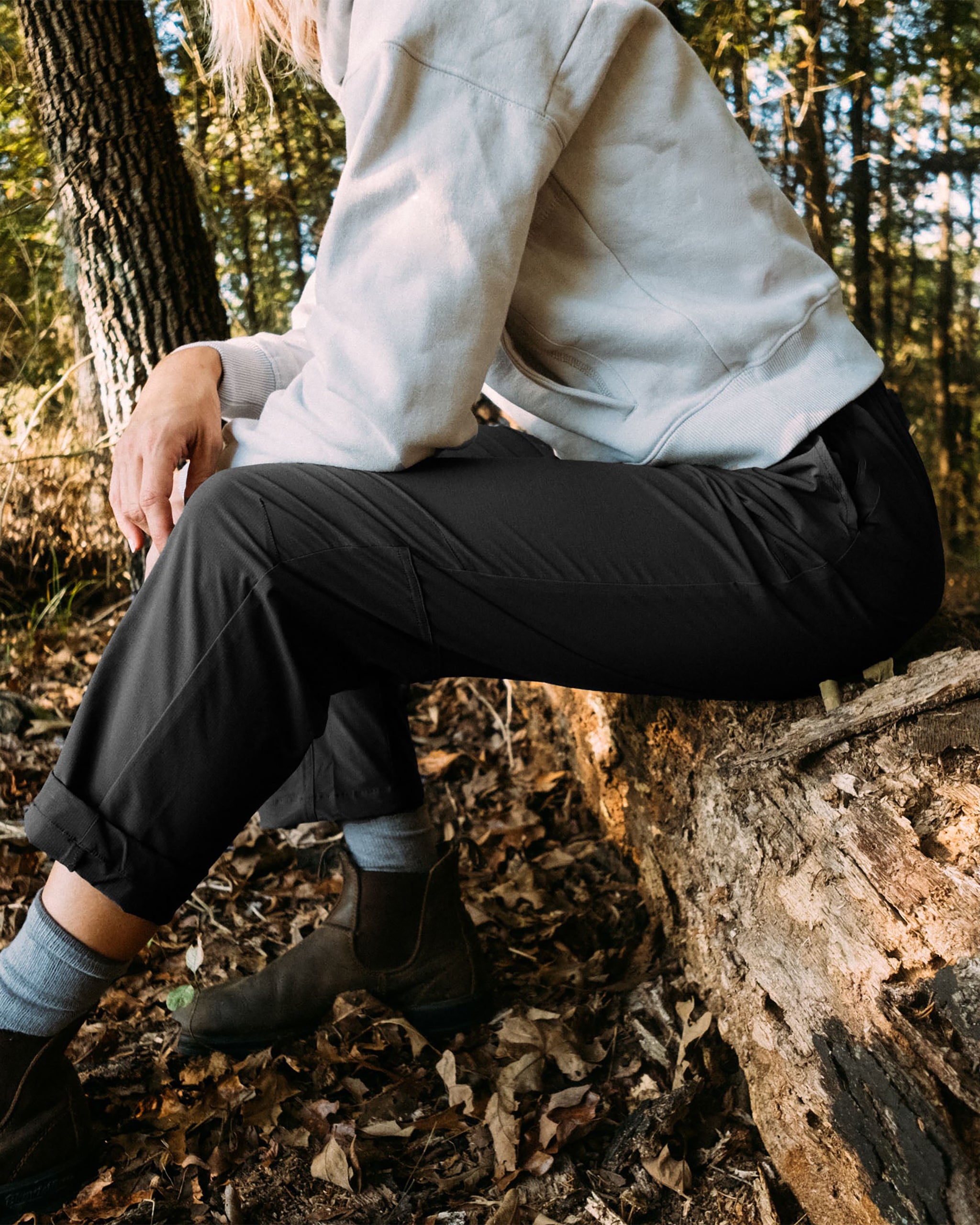 Wearing BEARDEDGOAT’s NEW — Summit™ Pant, a person sits on a fallen log amid autumn leaves and trees, their face hidden.