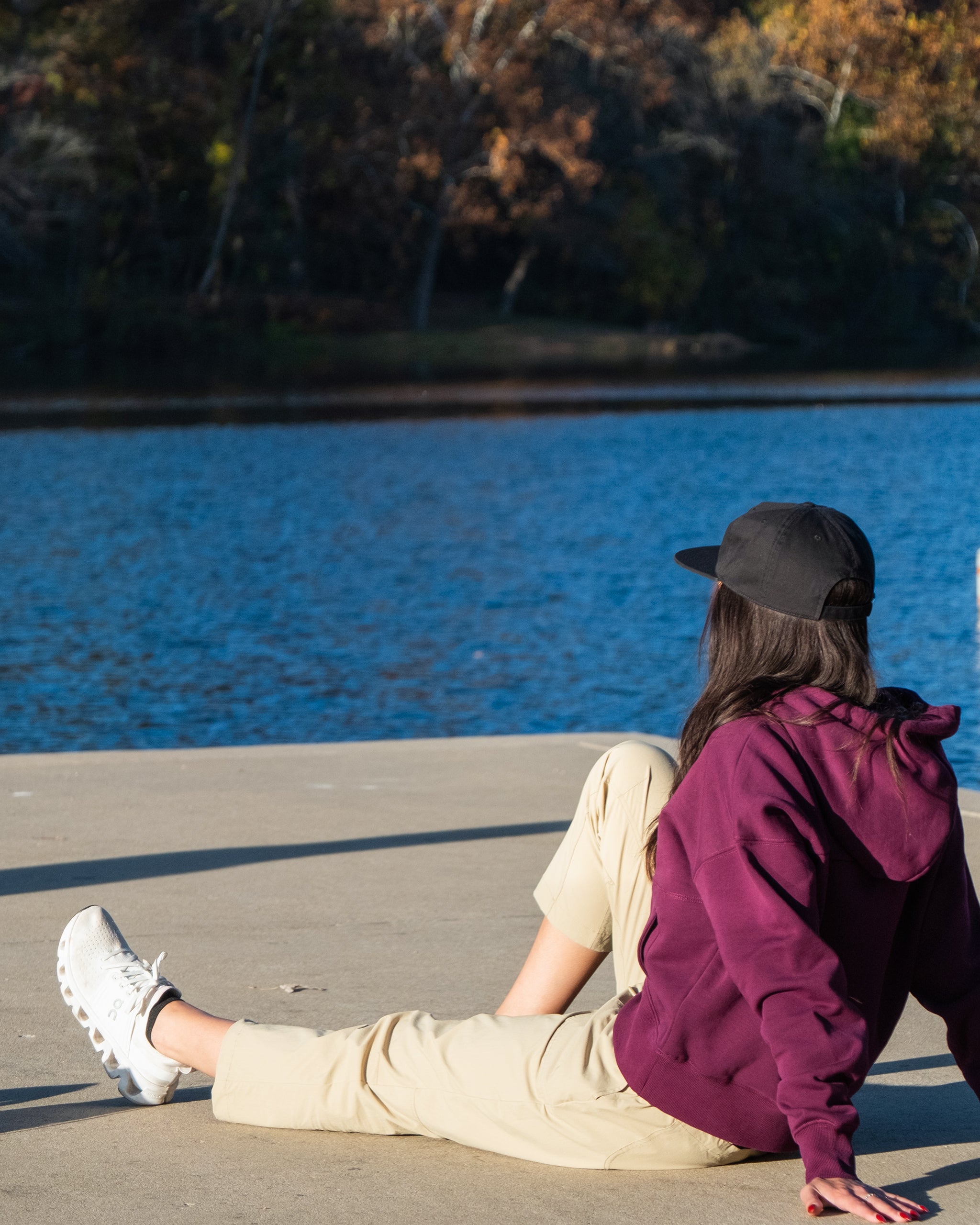 Wearing a maroon hoodie, BEARDEDGOAT’s NEW Summit™ Pant, white sneakers, and a black cap, a person sits on a concrete surface by a lake, gazing at the water with trees in the background.