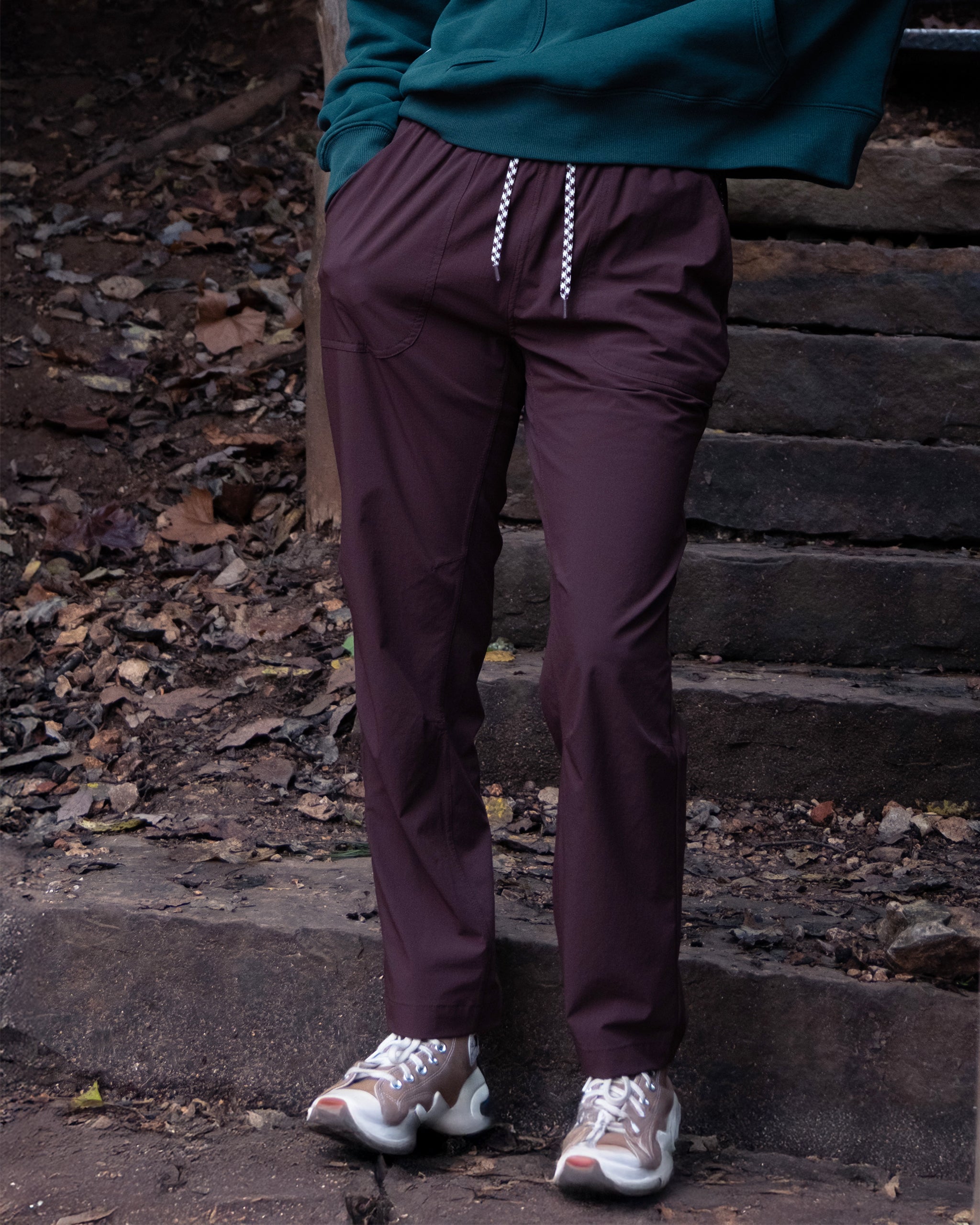 A person wearing maroon BEARDEDGOAT NEW — Summit™ Pant stands outdoors on stone steps with fallen leaves and dirt. They also wear a green hoodie and white sneakers; their upper body is not visible.