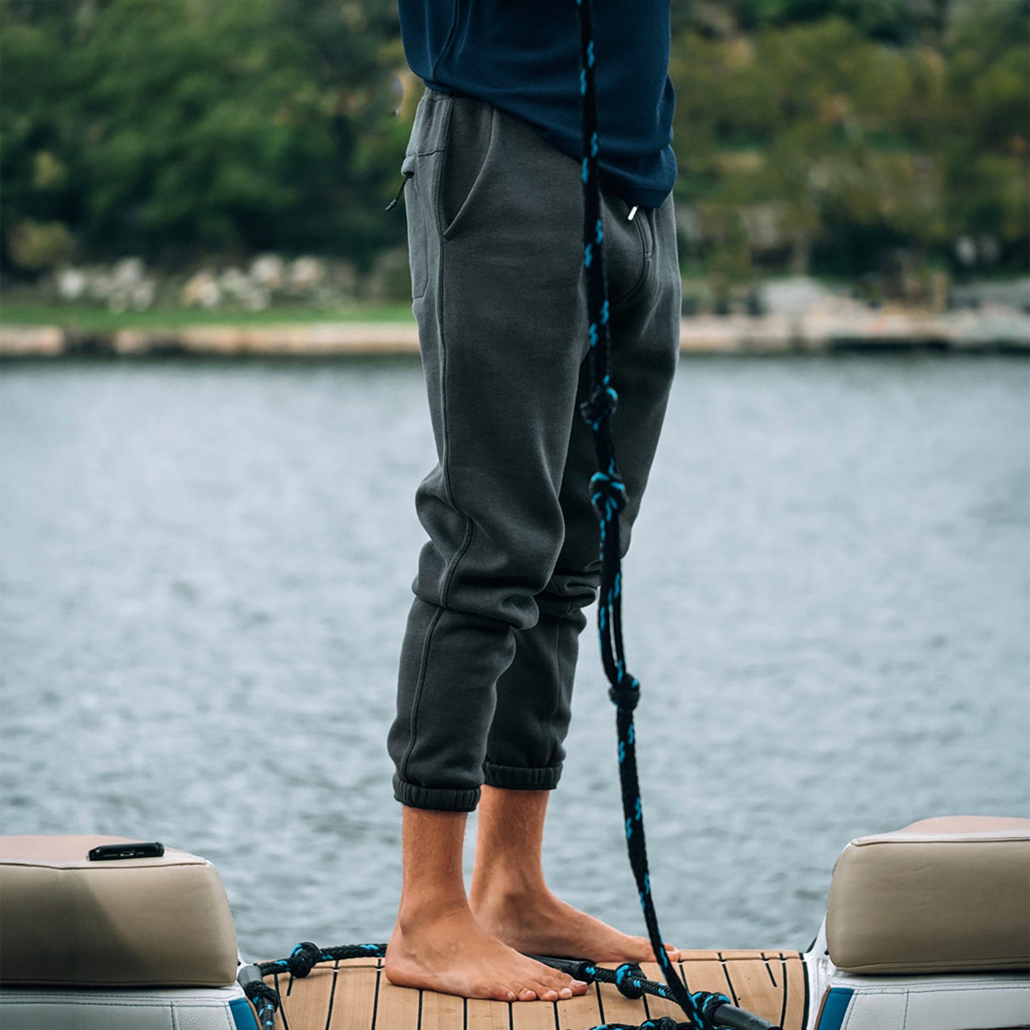 A person stands barefoot on a boat deck, wearing beardedgoat’s VOID™ Sweatpant and a long-sleeve top, with water and greenery in the background.