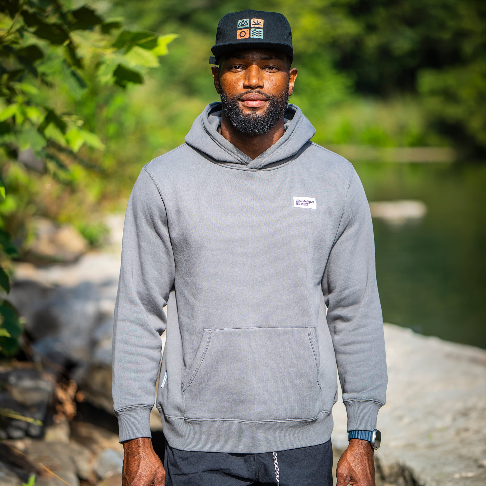 A man wearing The VOID™ Hoodie by beardedgoat and a black cap stands outdoors on a rocky path with greenery and water in the background. This premium heavyweight cotton fleece delivers warmth and style.