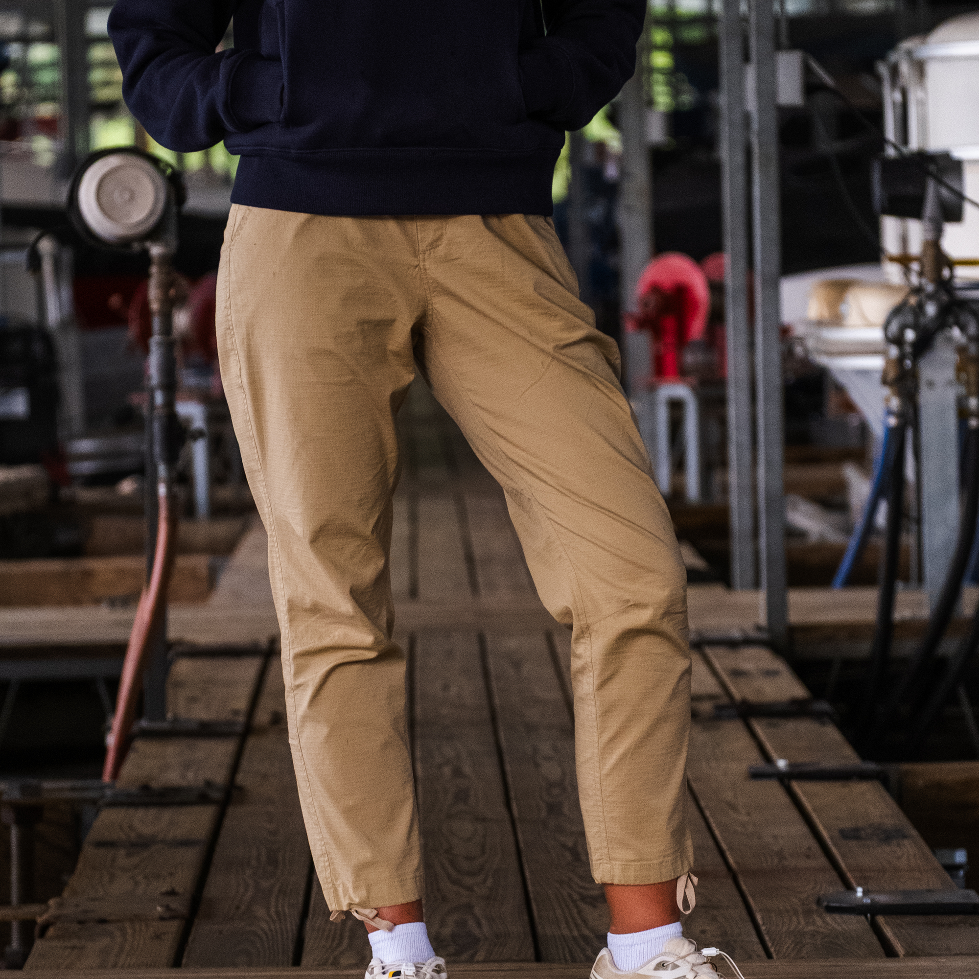 Wearing a dark hoodie, BEARDEDGOAT Women's Canopy Pant, and sneakers, a person stands with hands in their pockets on a wooden dock with industrial equipment and pipes in the background.