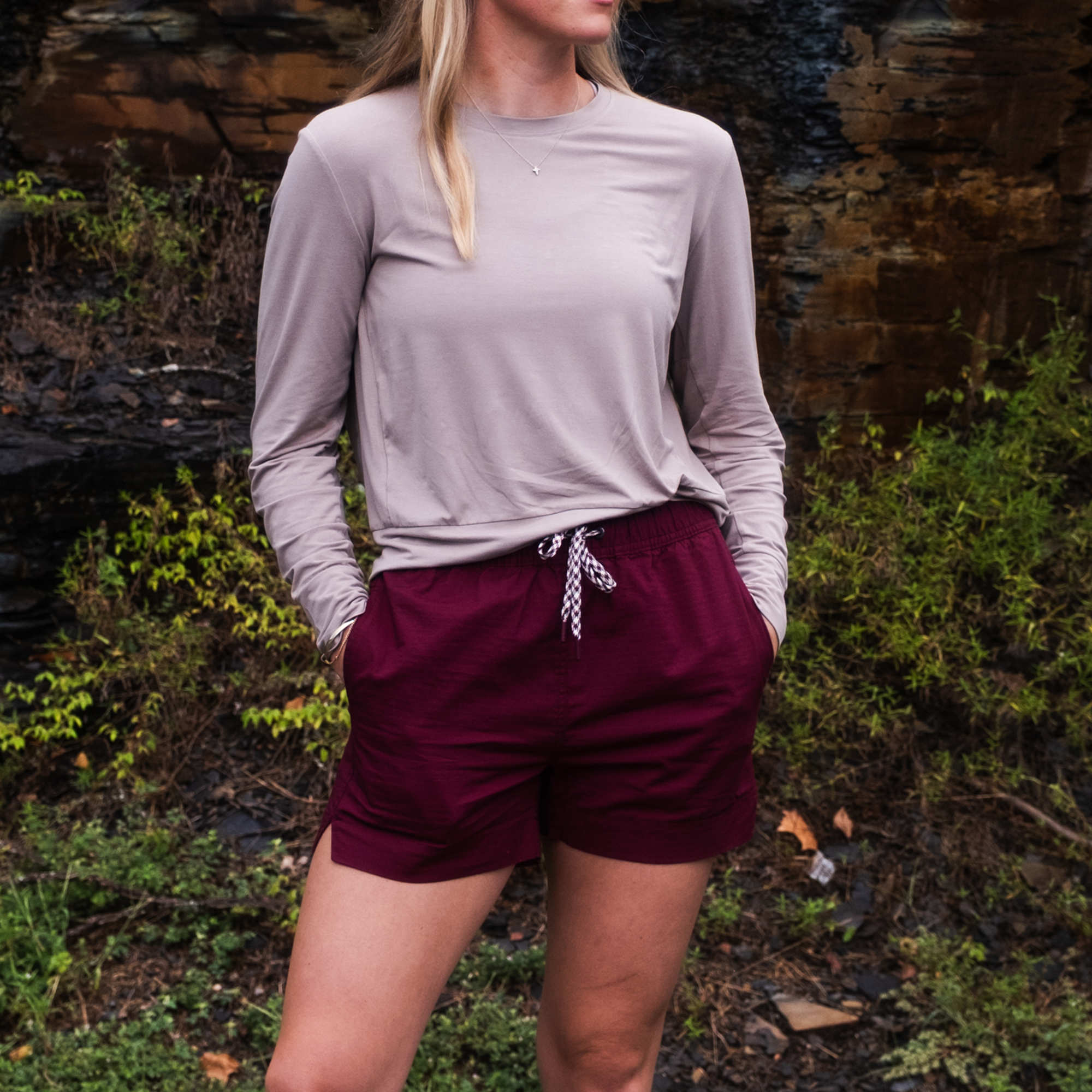 Outdoors among rocks and green plants, a person with hands in pockets wears BEARDEDGOAT's Women's Canopy Short and a long-sleeve beige shirt. Their face is partially obscured.