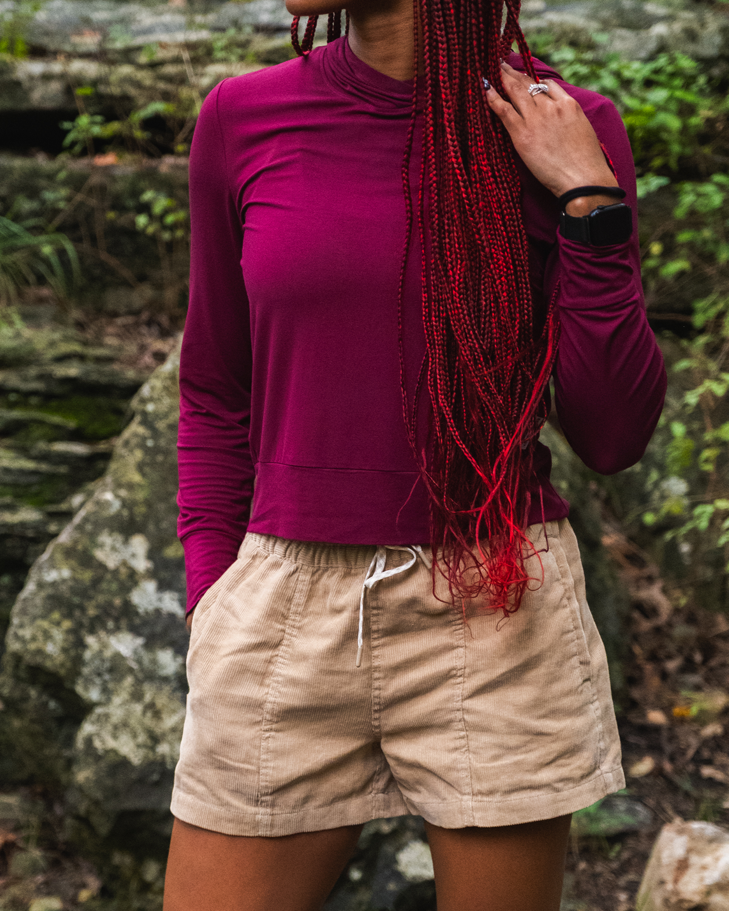 A person with long red braids, wearing a maroon long-sleeve shirt and BEARDEDGOAT Women's Cord Shorts in beige, stands outdoors before large rocks and greenery. Their face is partially out of frame.