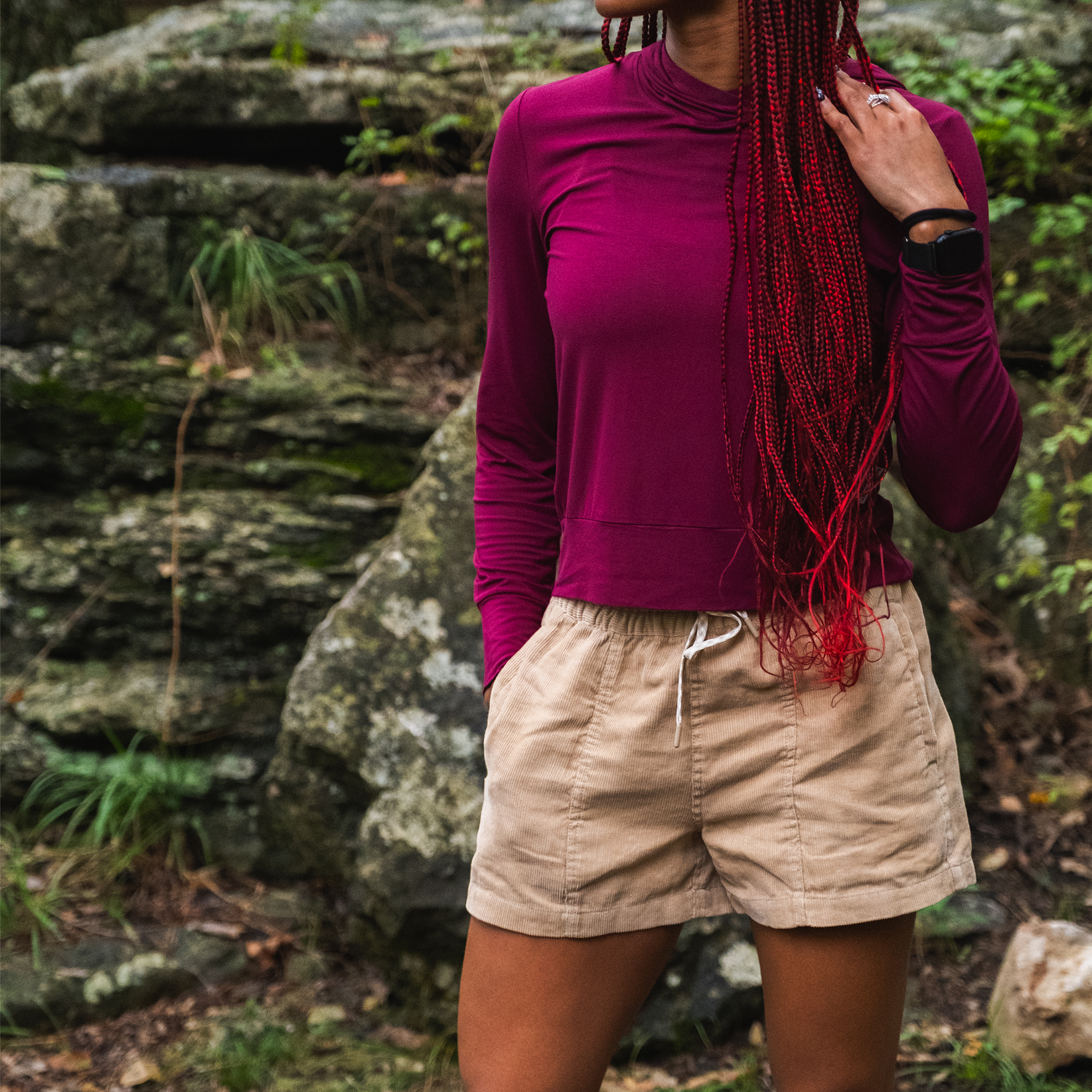 A person with long red braids, wearing a maroon long-sleeve shirt and BEARDEDGOAT Women's Cord Shorts in beige, stands outdoors before large rocks and greenery. Their face is partially out of frame.